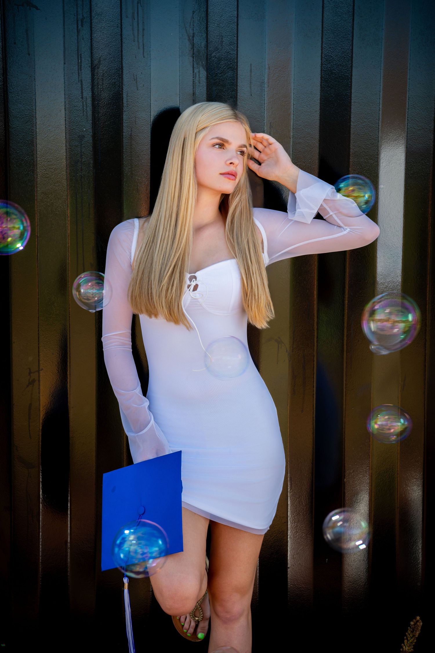Omaha Senior Photography - Photographer in Omaha - Model in white dress poses against dark fence with floating soap bubbles.