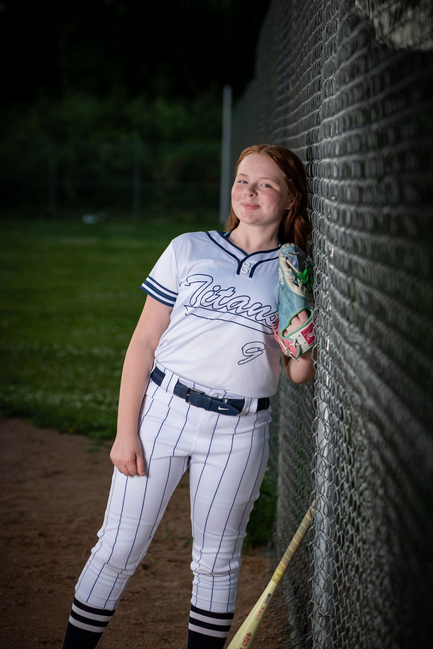 Softball player in white pinstriped uniform leans against chain link fence with mitt and green background during sports photography session.