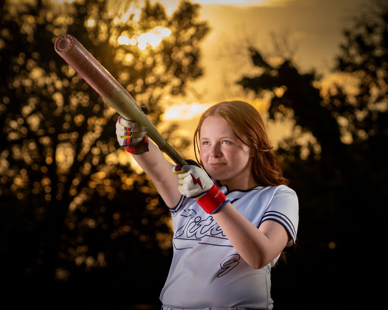 Youth softball player holds a bat against a dramatic sunset backdrop with trees silhouetted in the background during Omaha sports session.