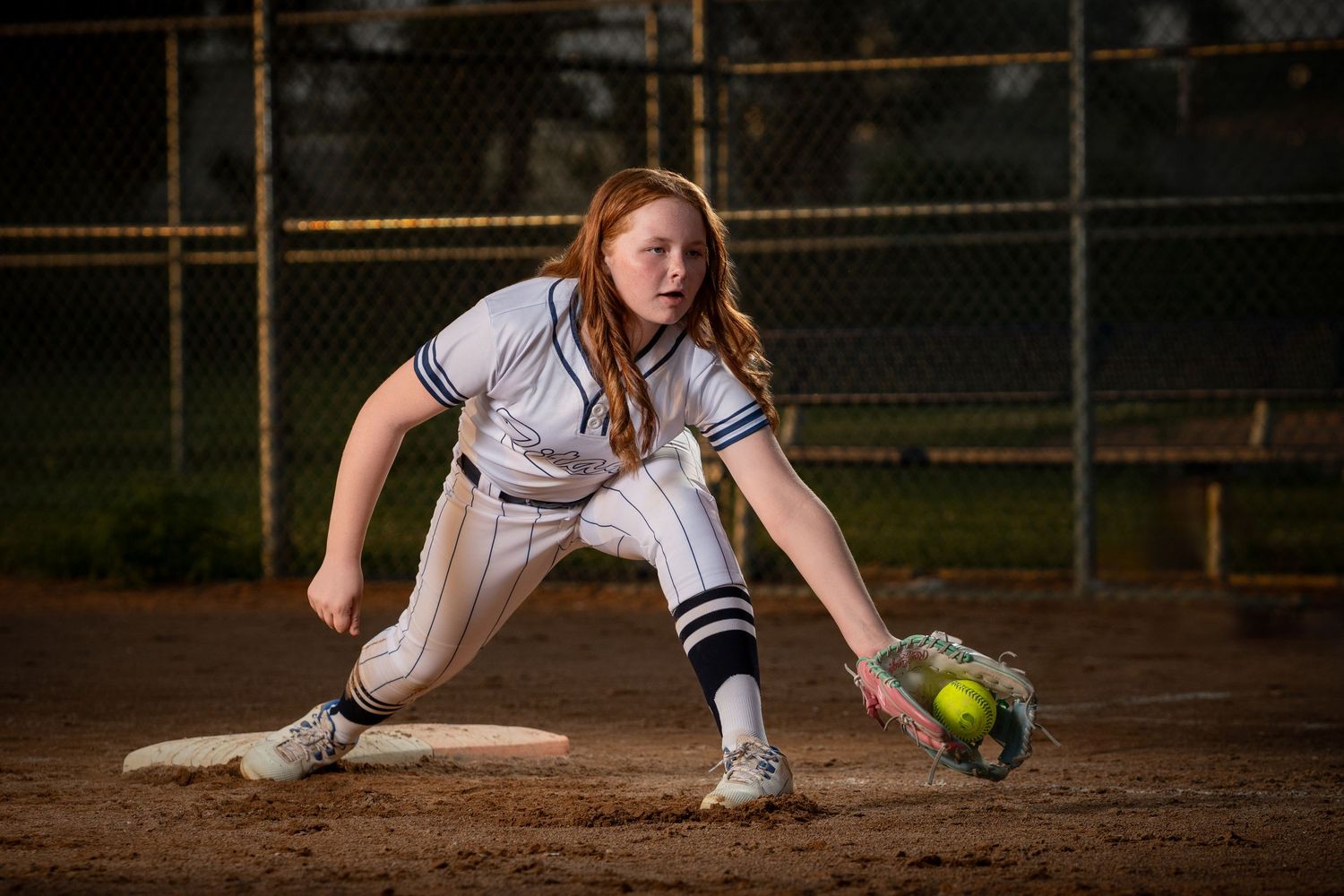 Softball player in white uniform stretches to field a ground ball on dirt infield during evening game taken by Omaha sports photographer.