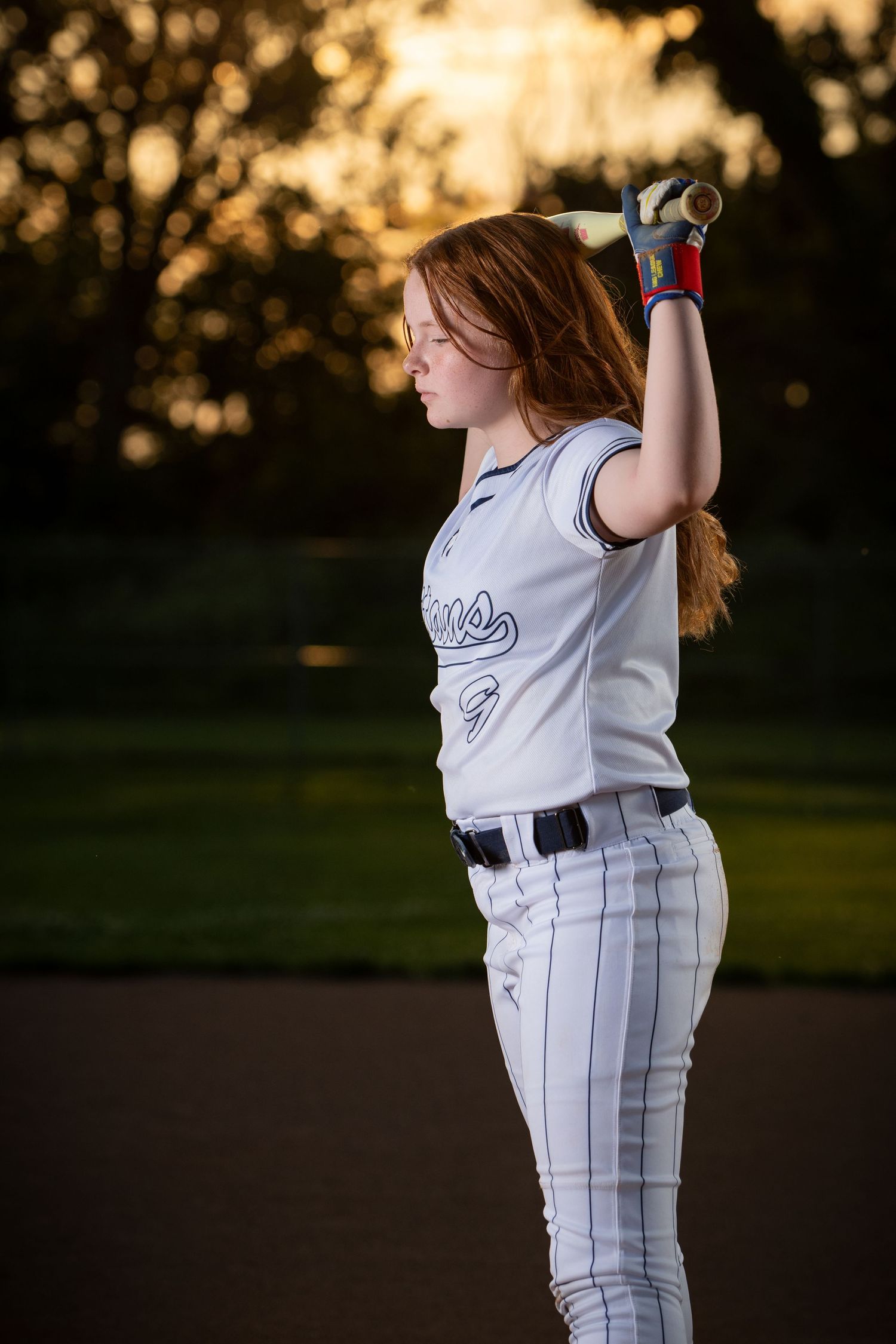 Softball player in white pinstriped uniform stands in golden sunset light practicing batting stance on field by Iowa sports photographer.