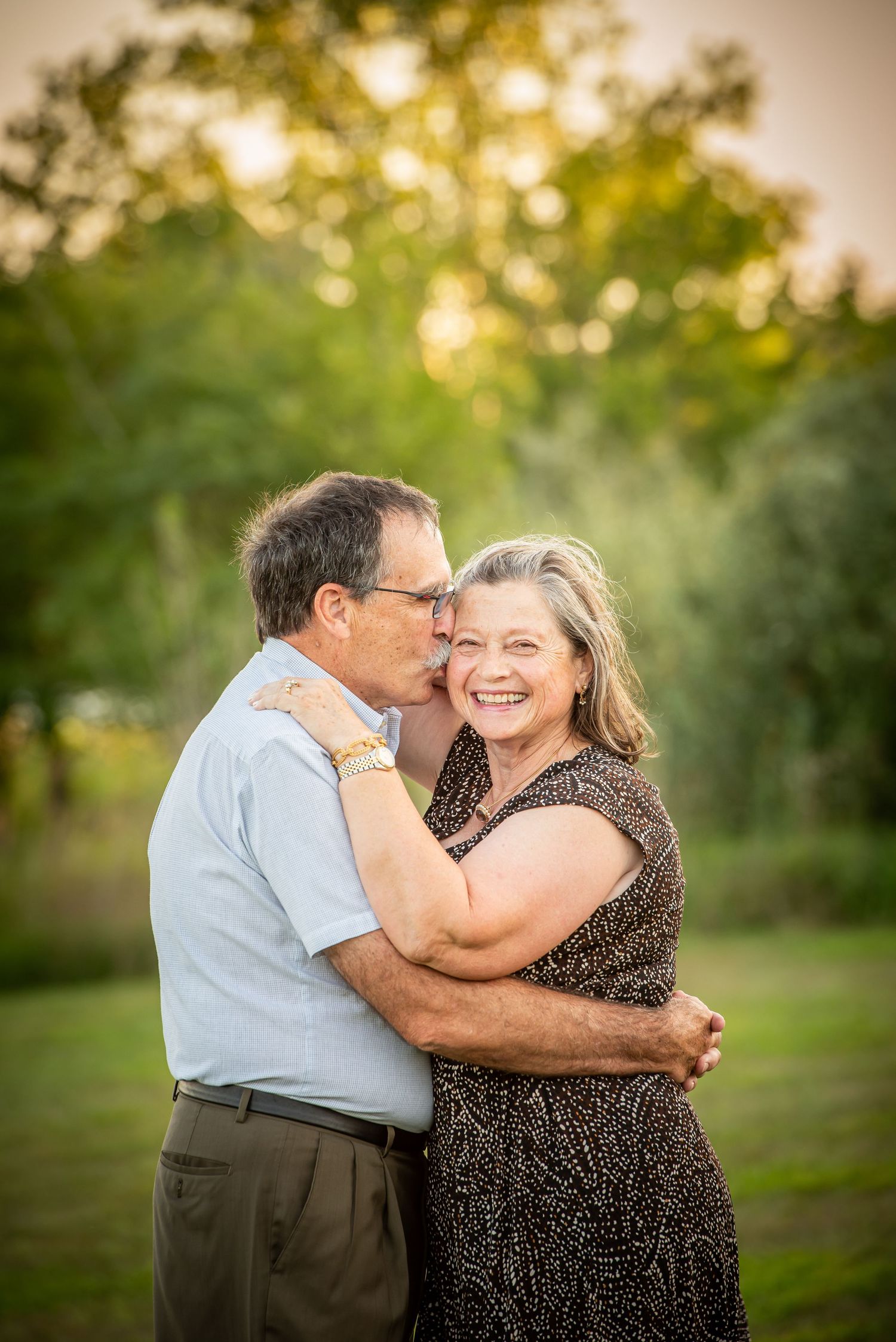 Chalco Lake Family Photography - An affectionate couple embrace and smile in a sunny outdoor setting with bokeh lights and greenery.