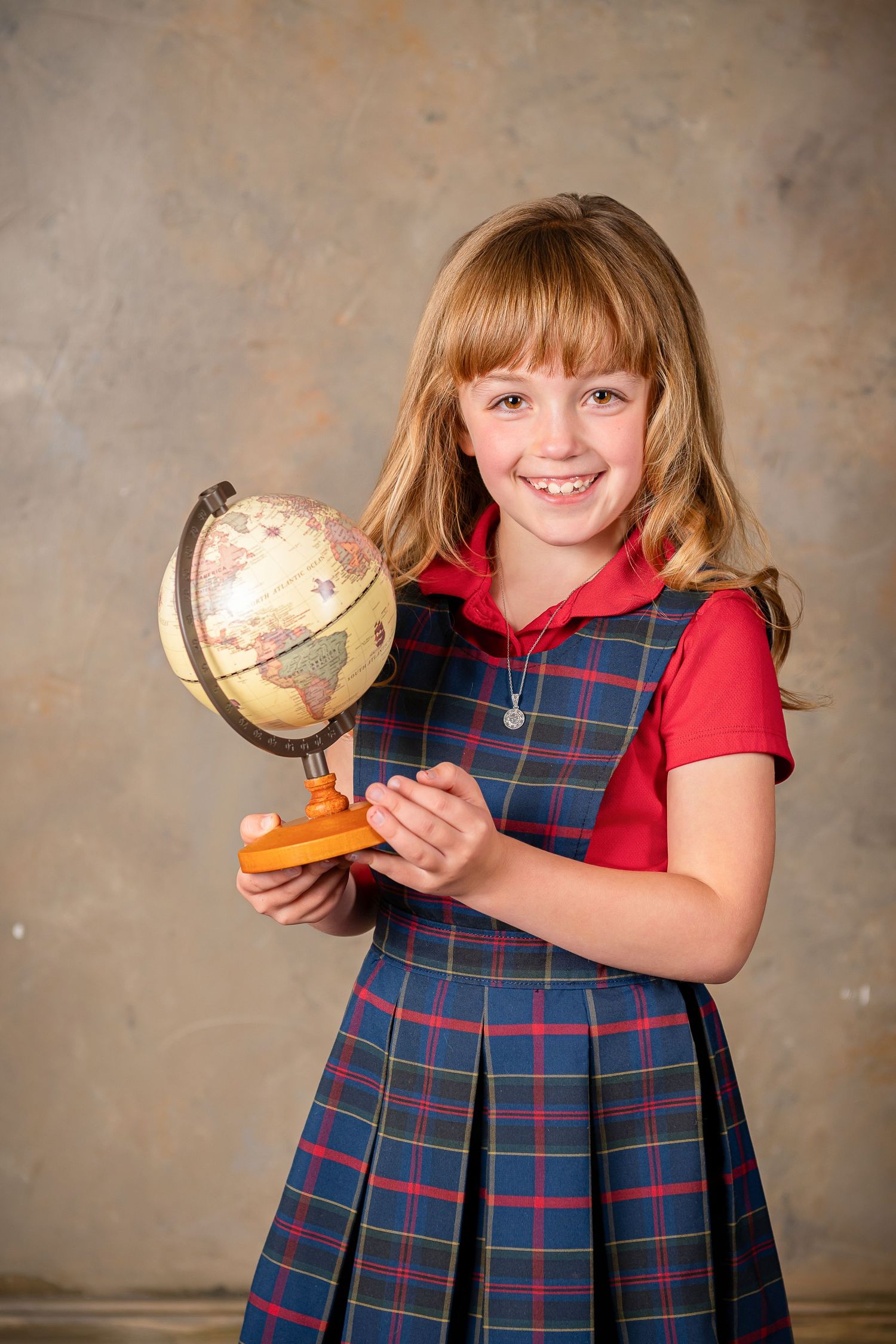 Student in plaid uniform dress and red shirt holds small decorative globe while posing for school photo for children's photo session Omaha.