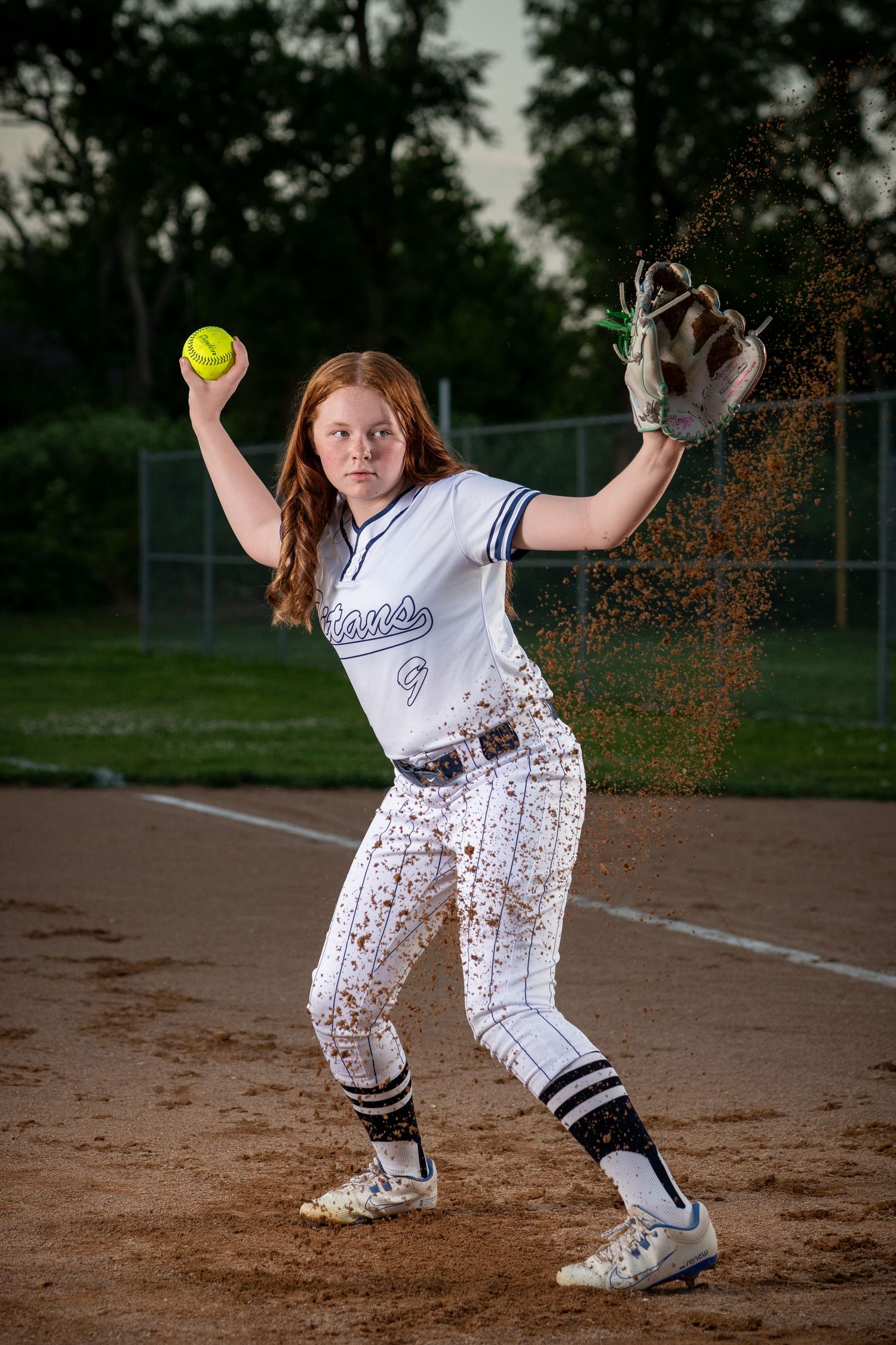 Softball player in white uniform pitches on a dirt infield at dusk with a dramatic dirt splash around them by Iowa sports photographer.