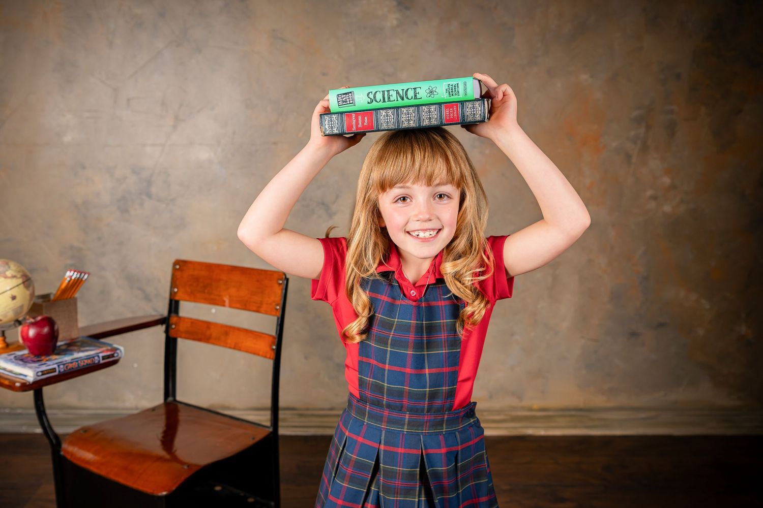 A young student in a plaid uniform dress holds a green toy ruler above their head while smiling with Omaha children's photographer.
