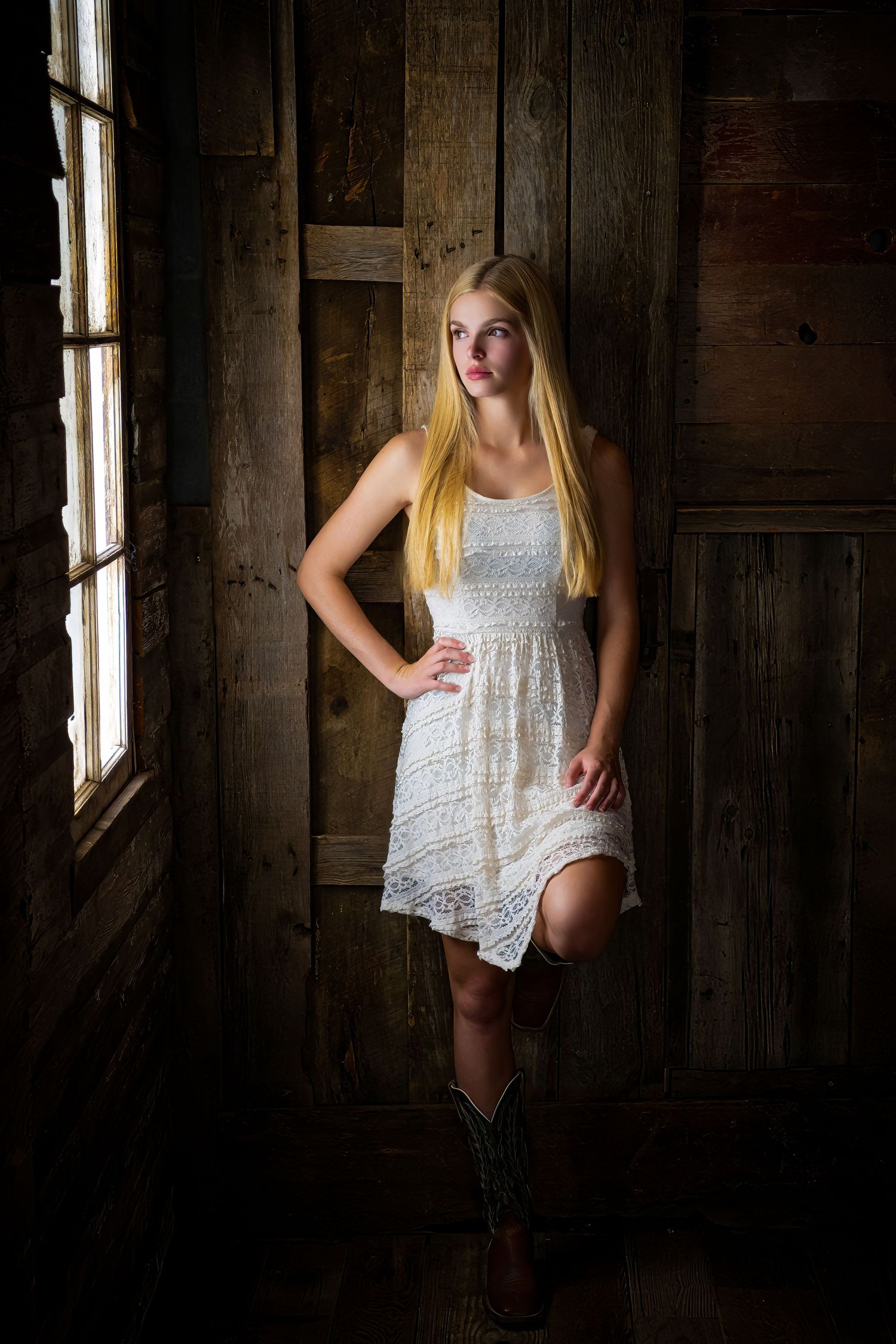 Omaha Senior Photography - Photographer in OmahaCountry style portrait of woman in white lace dress and cowboy boots in rustic barn setting.