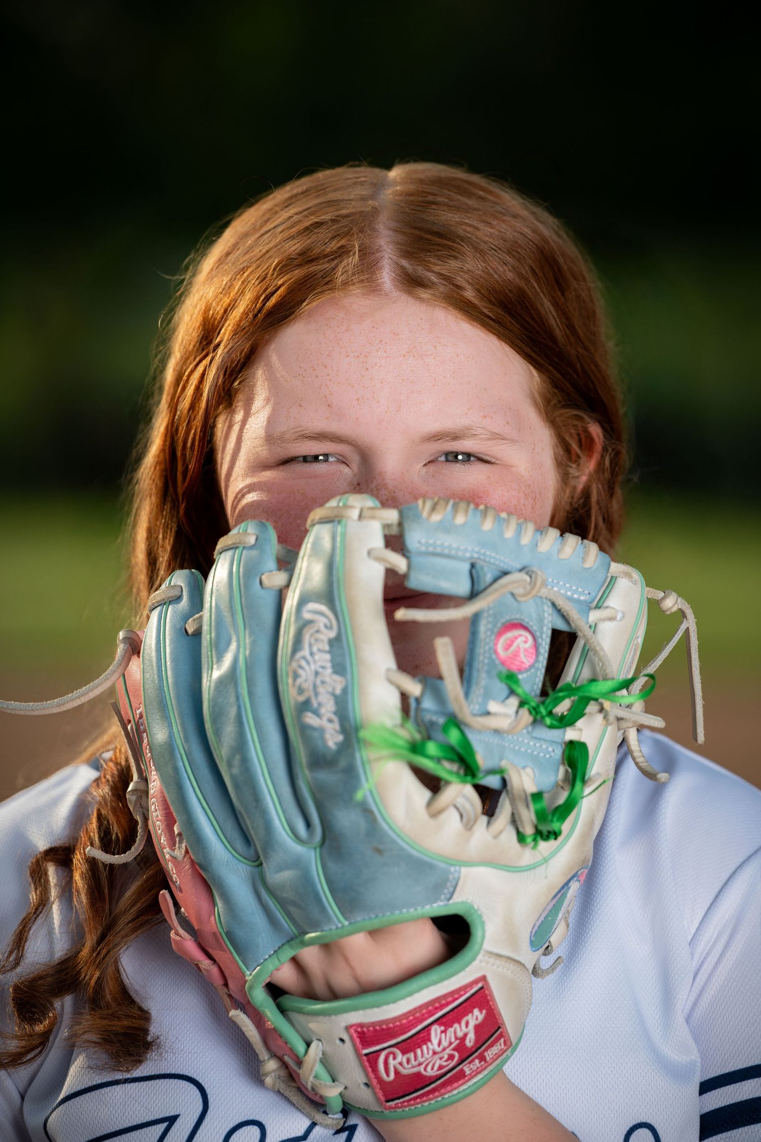 A softball player in a white uniform holds up a light blue and white Rawlings baseball glove near their face during sports photo session.