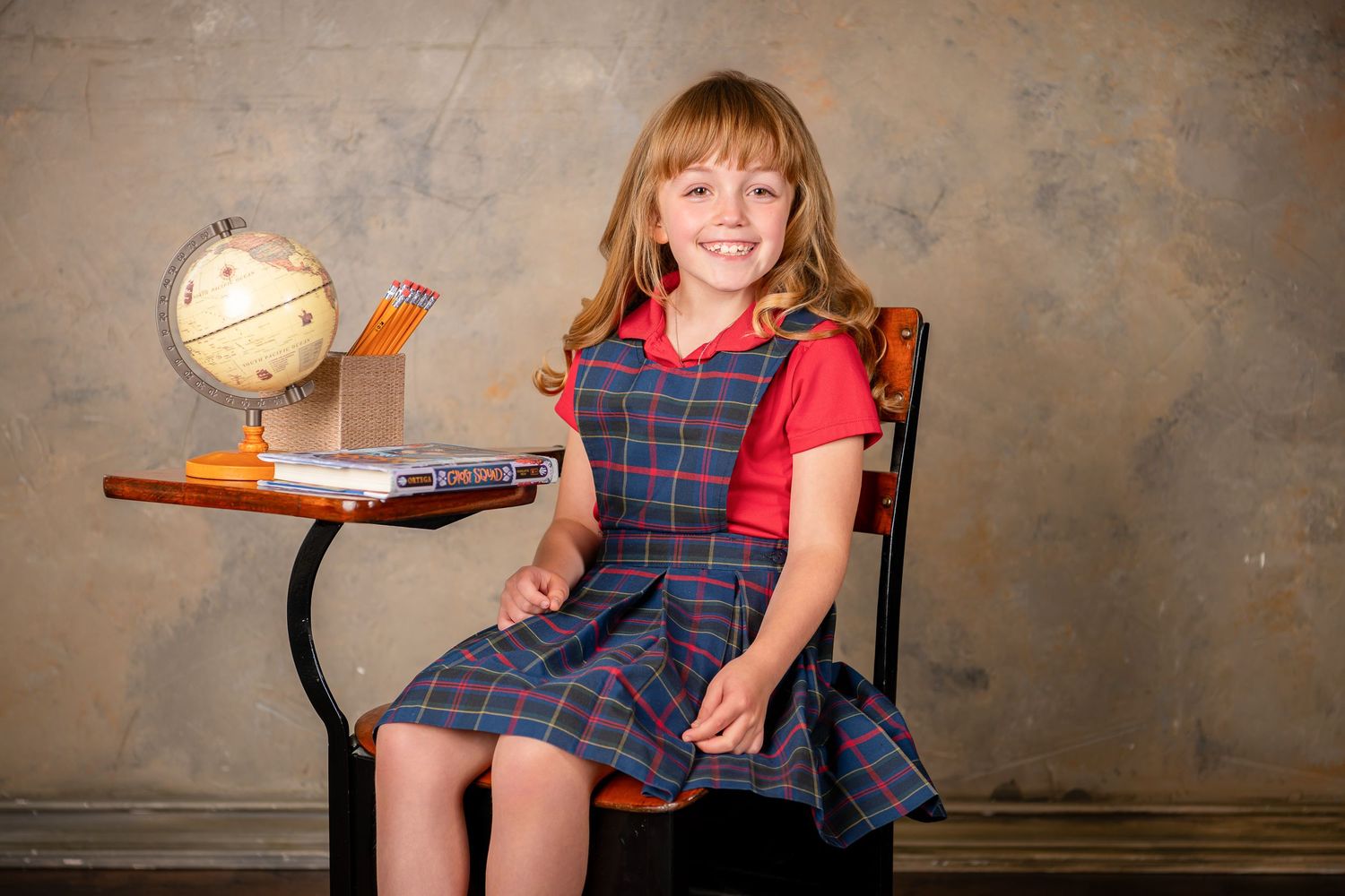 A student in a plaid skirt uniform sits on a wooden chair outdoors against a textured wall during Omaha back to school photography session.