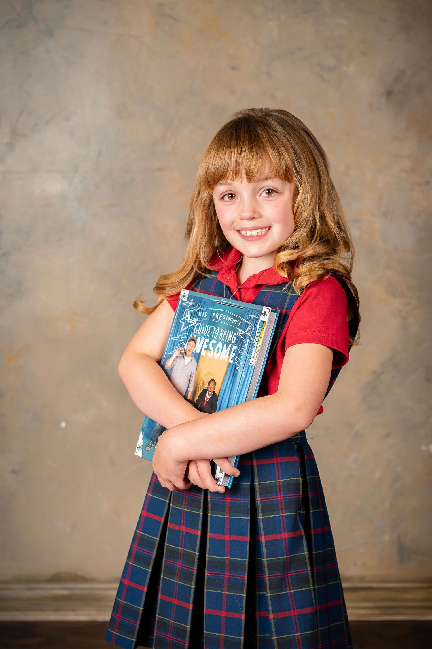 Student wearing plaid uniform holding textbook against textured backdrop in studio portrait take by Omaha children's photographer.