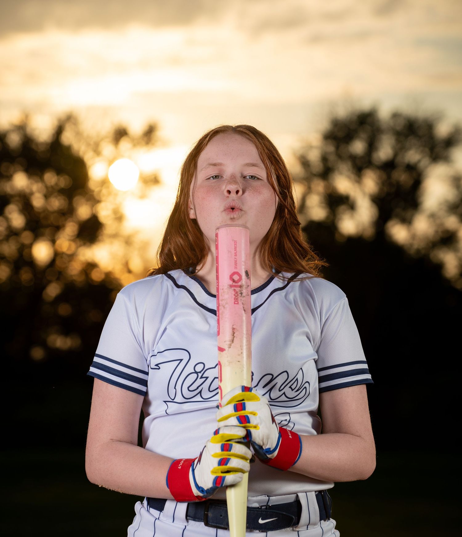 Softball player in white Tigers jersey holds up pink popsicle against golden sunset backdrop during Council Bluffs sports photo session.