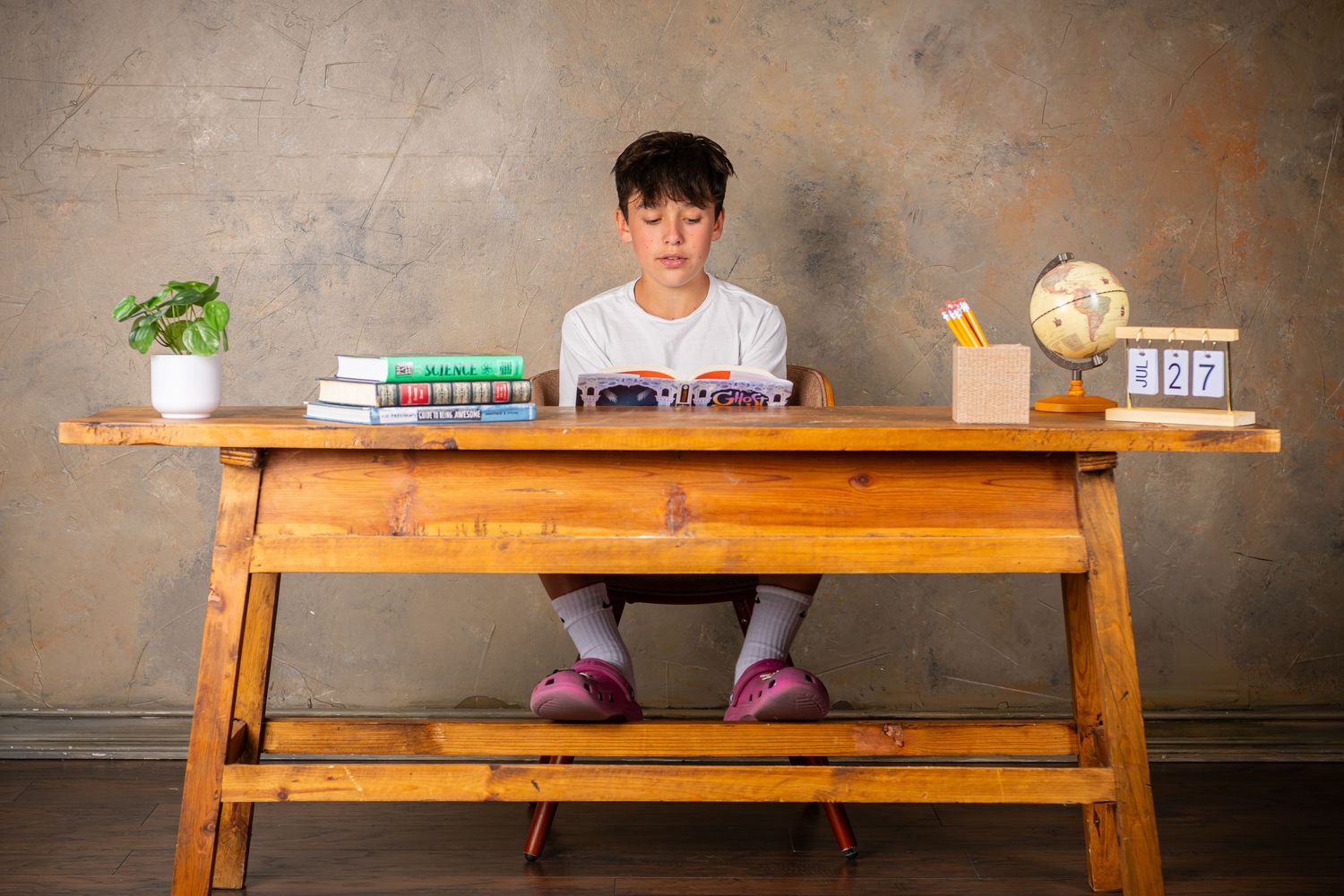 A wooden desk is decorated with books, a plant and decorative items against a neutral wall background during back to school photo session.