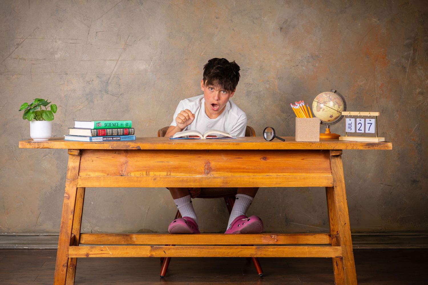 Student writing at wooden desk with books, globe and digital clock on textured gray wall background during child photography session Omaha.