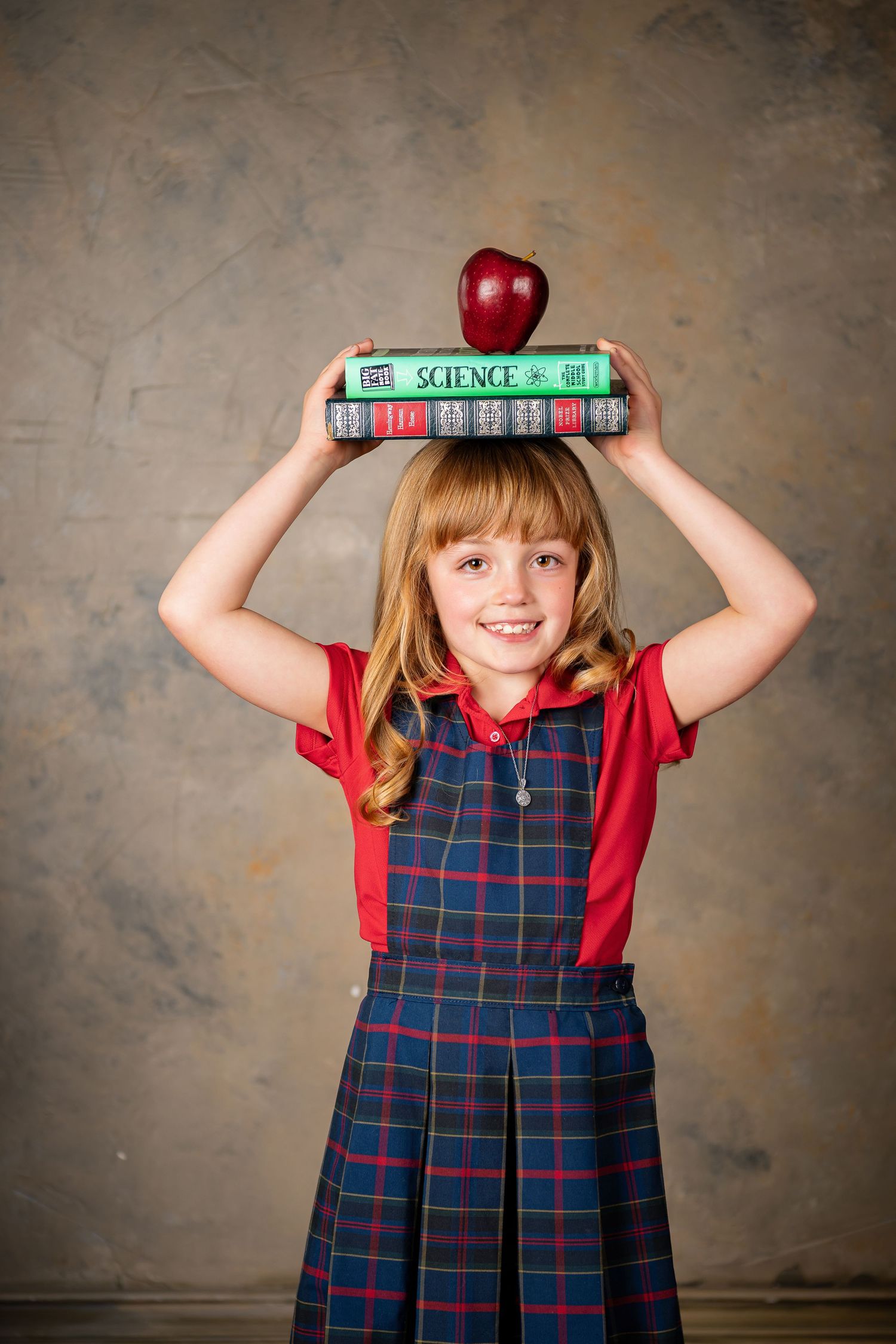 Student in plaid uniform balancing books and red apple on head against studio backdrop during Omaha children's photography session.