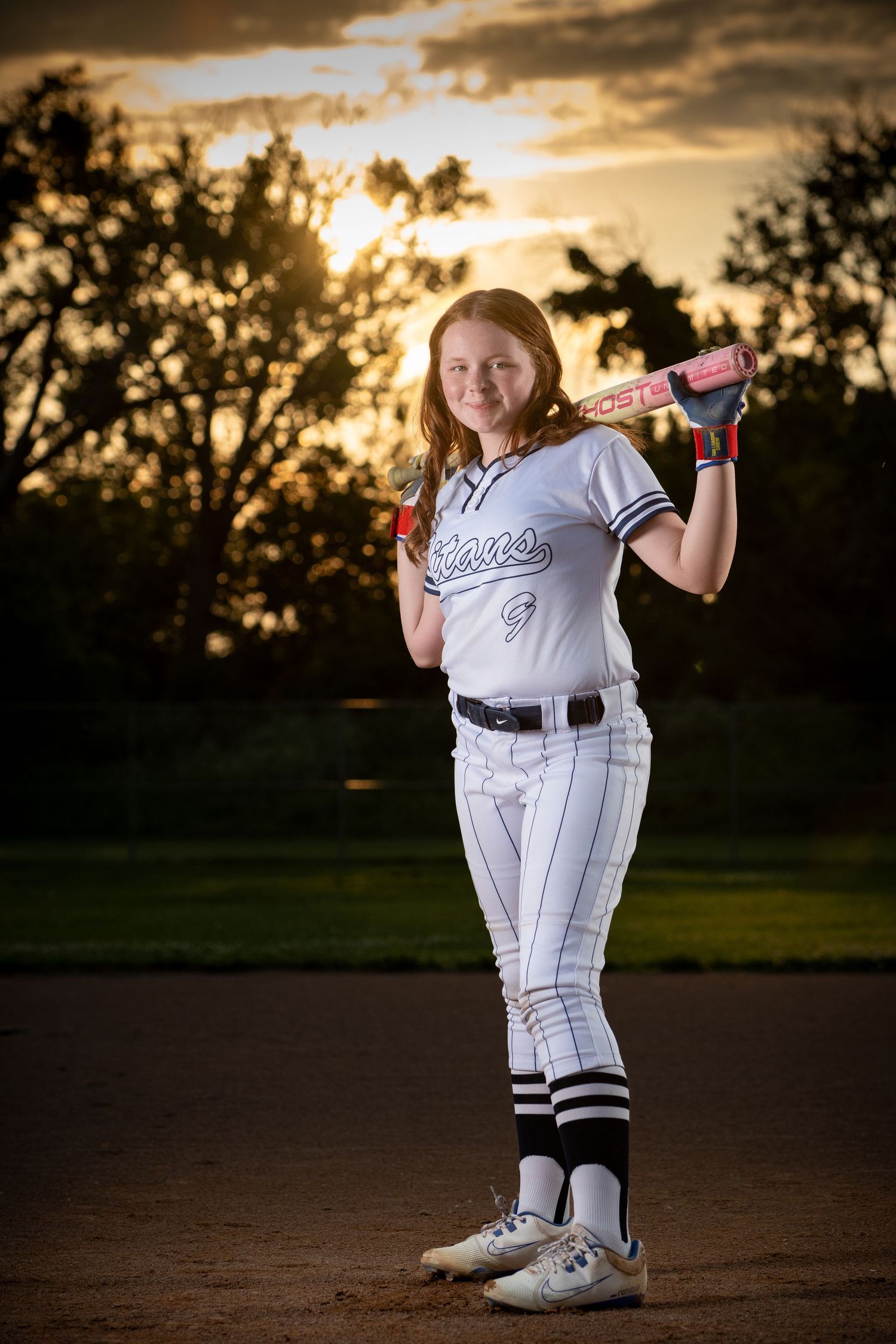 Youth softball player in white uniform poses with pink bat at sunset on baseball diamond as golden light filters through trees Omaha photos.