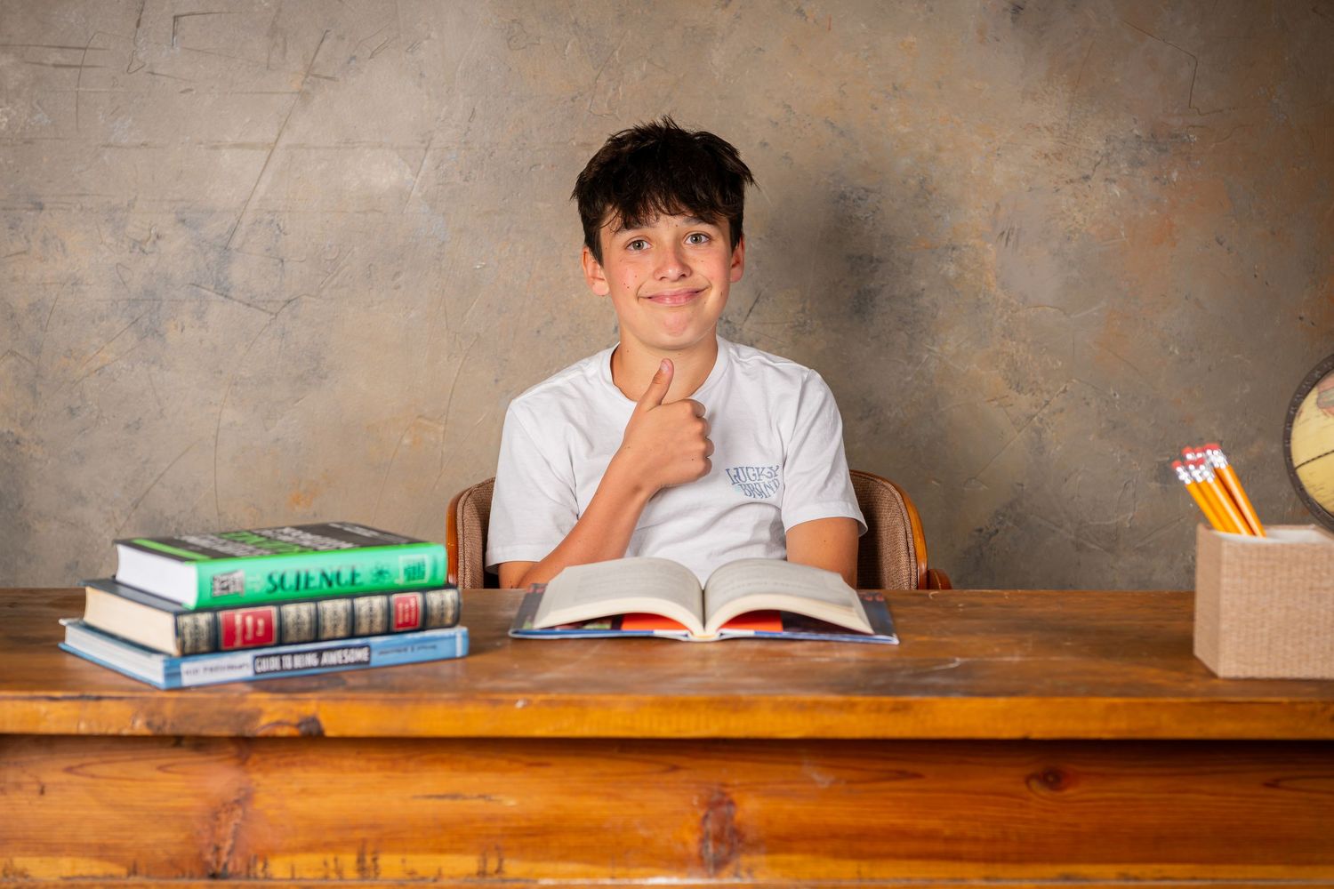 Student sits at wooden desk with textbooks and pencil holder while studying and smiling at camera during a back to school photo session.