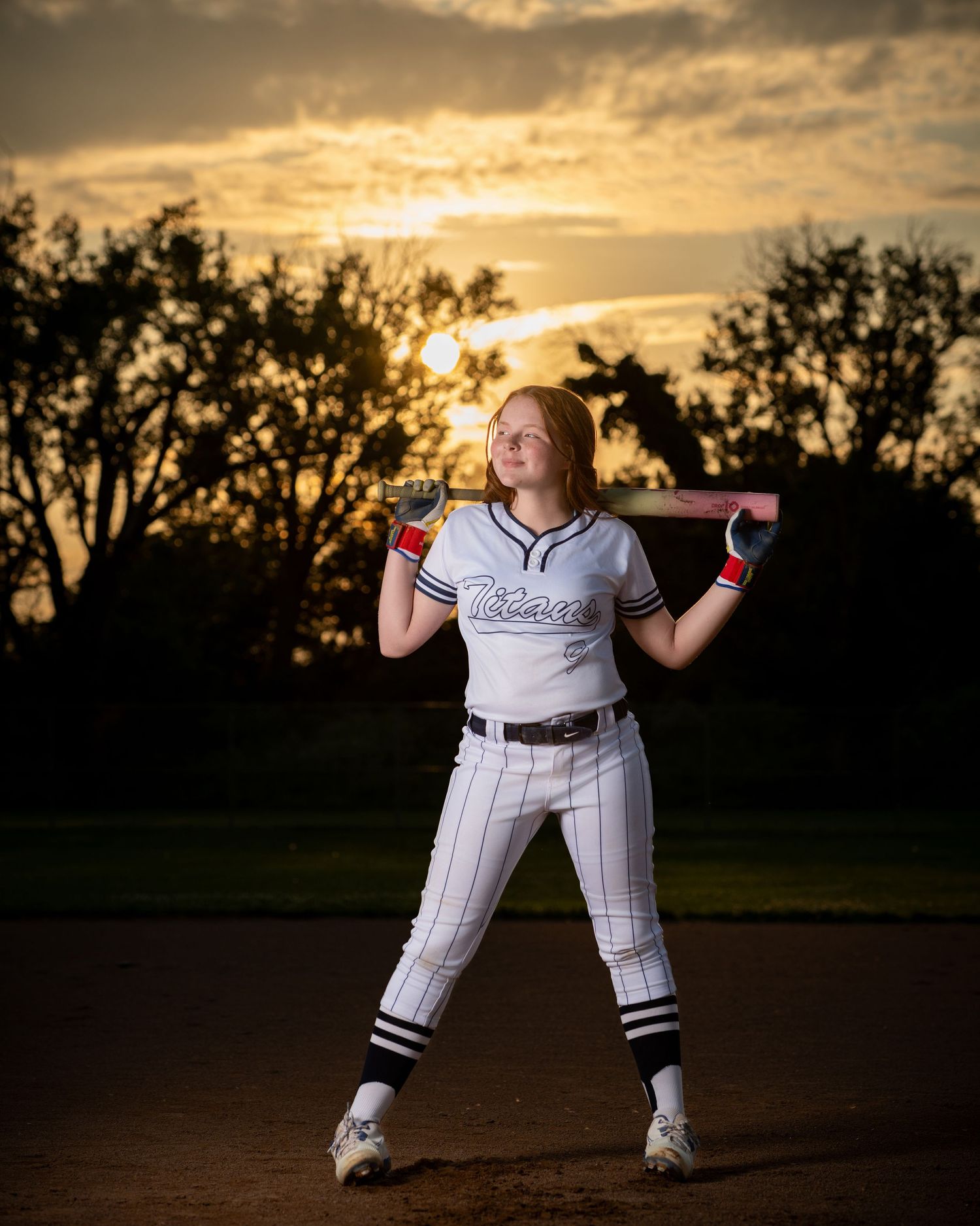 Baseball player in pinstriped uniform poses with bat at sunset on field with dramatic golden sky behind during Iowa sports photo session.