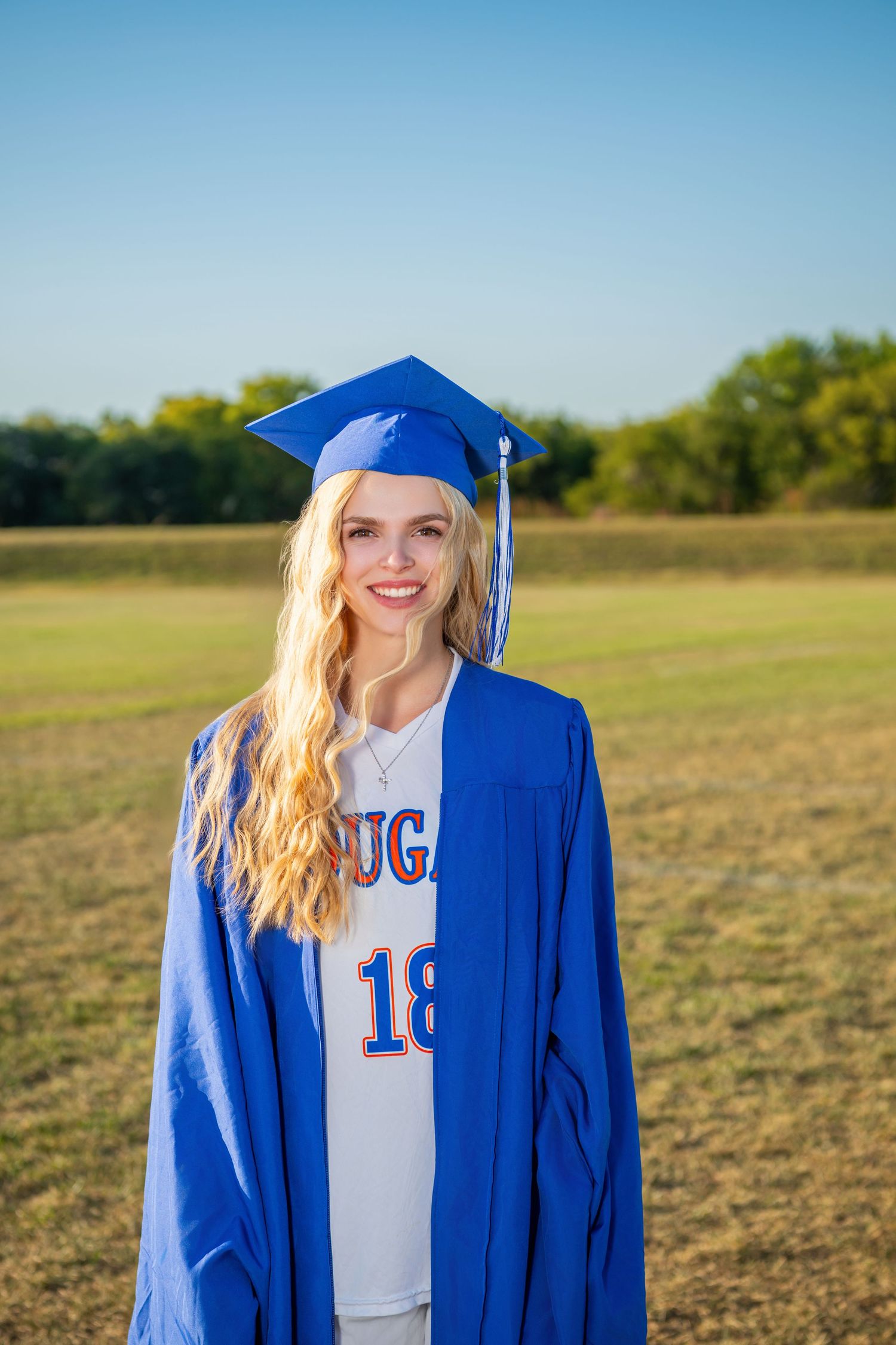 Omaha Senior Photography - Photographer in Omaha - Graduate in blue cap and gown stands on athletic field during golden hour.