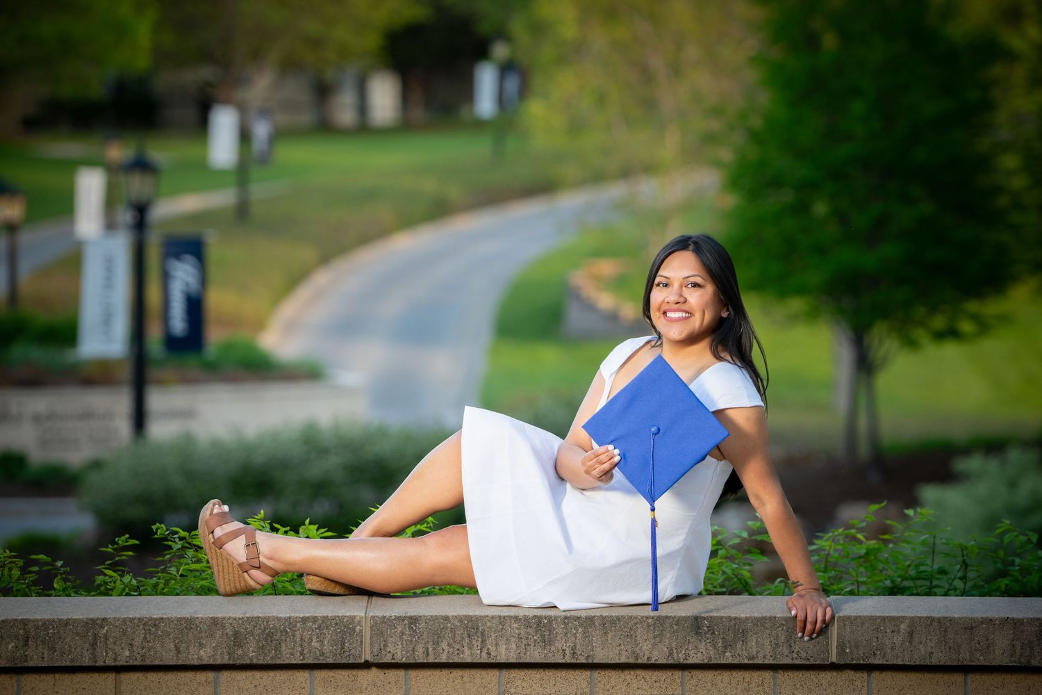 Graduation photographer in Omaha - A student in a white dress sits on stone steps outdoors holding a blue folder in a scenic park setting.