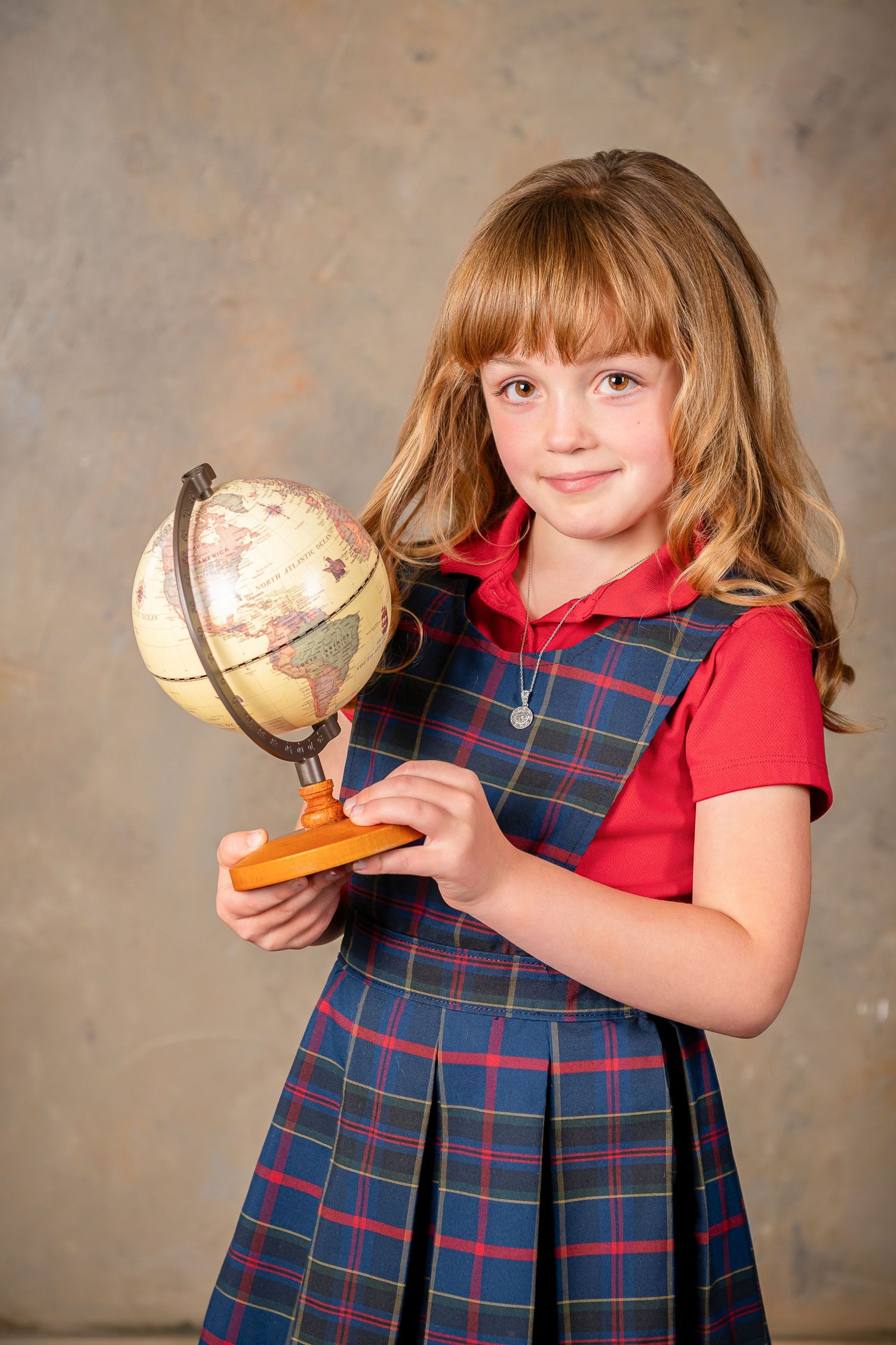 Girl in red and plaid uniform poses with vintage globe against textured wall during a back to school photo session in Omaha, Nebraska.