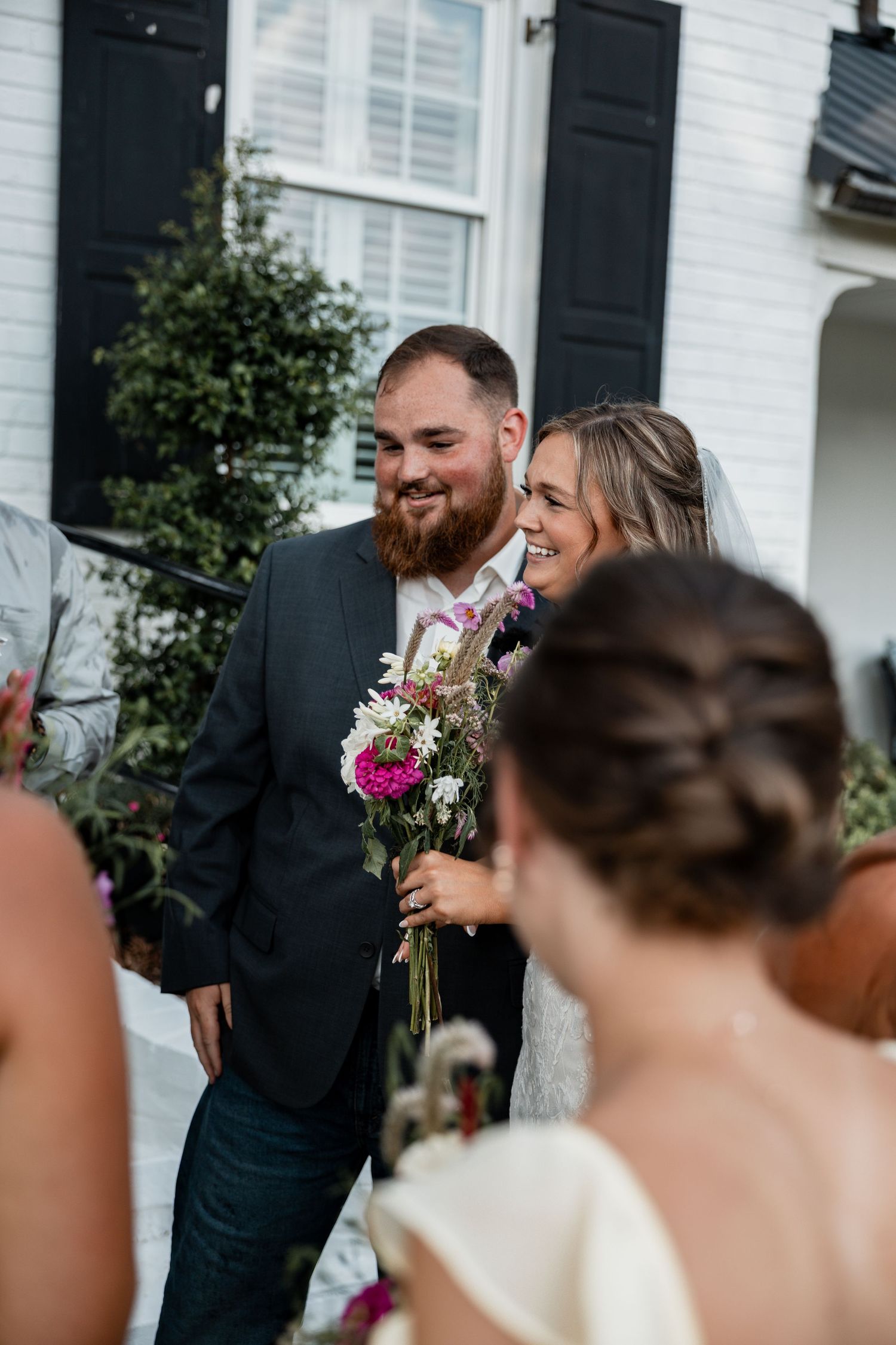 Wedding couple sharing a joyful moment outside white house with black shutters.