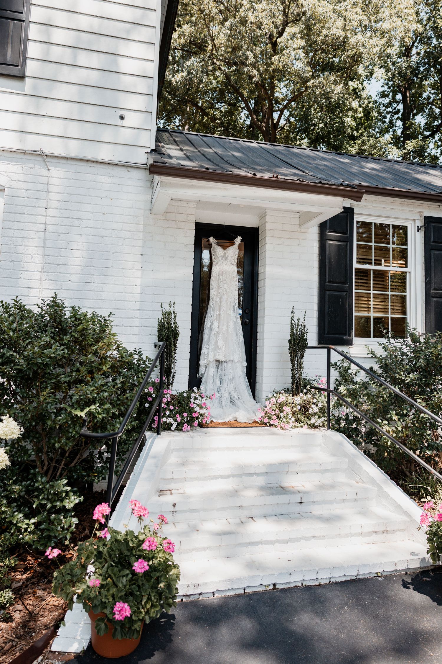 White wedding dress hanging in doorway of classic white house with black shutters and pink flowers.