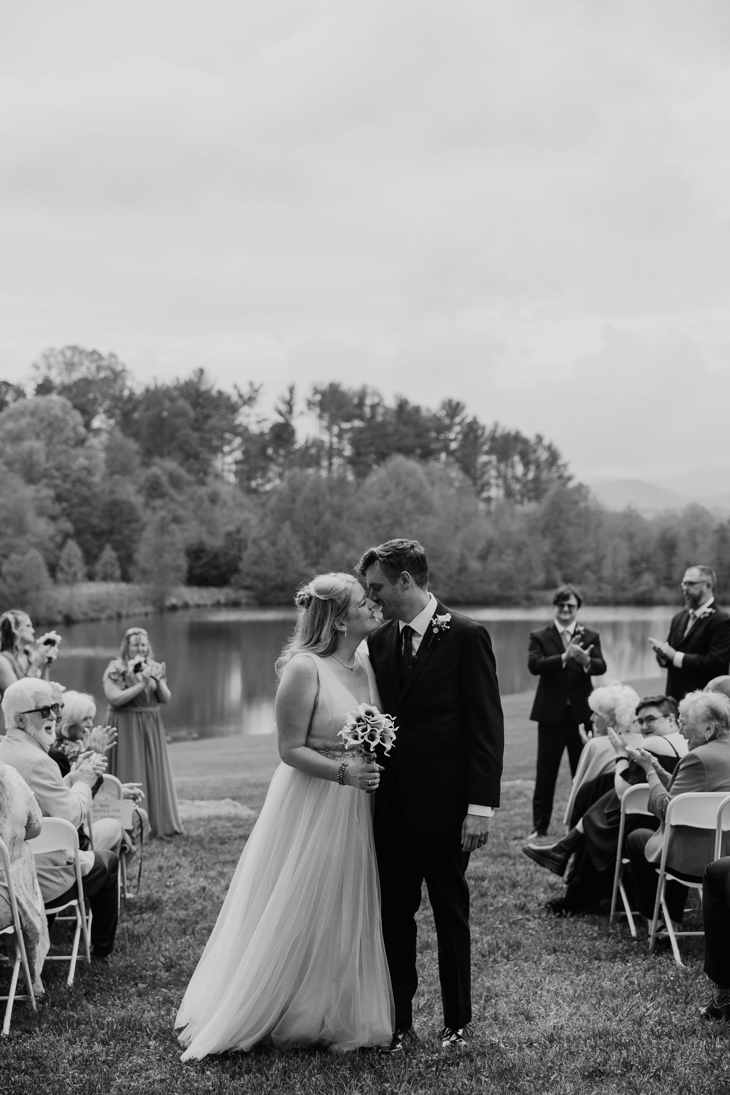 Black and white photo of wedding ceremony by lake with guests seated on folding chairs.