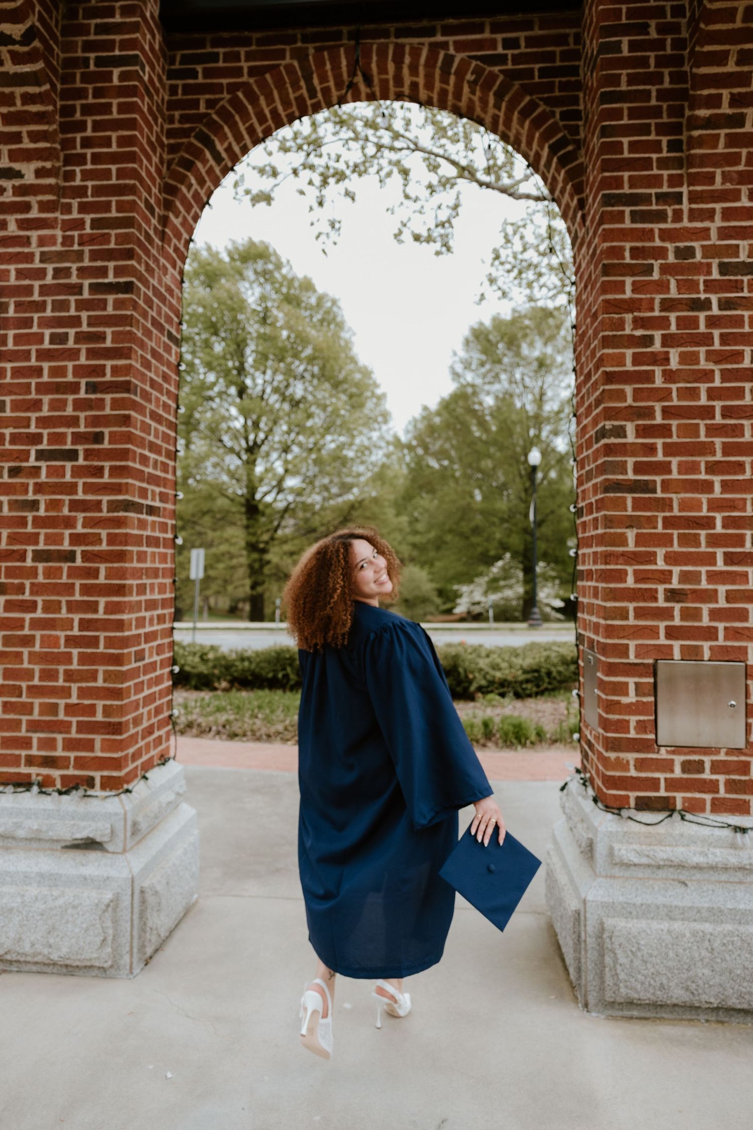 Graduate in blue cap and gown twirls joyfully under a brick archway during graduation photo session.