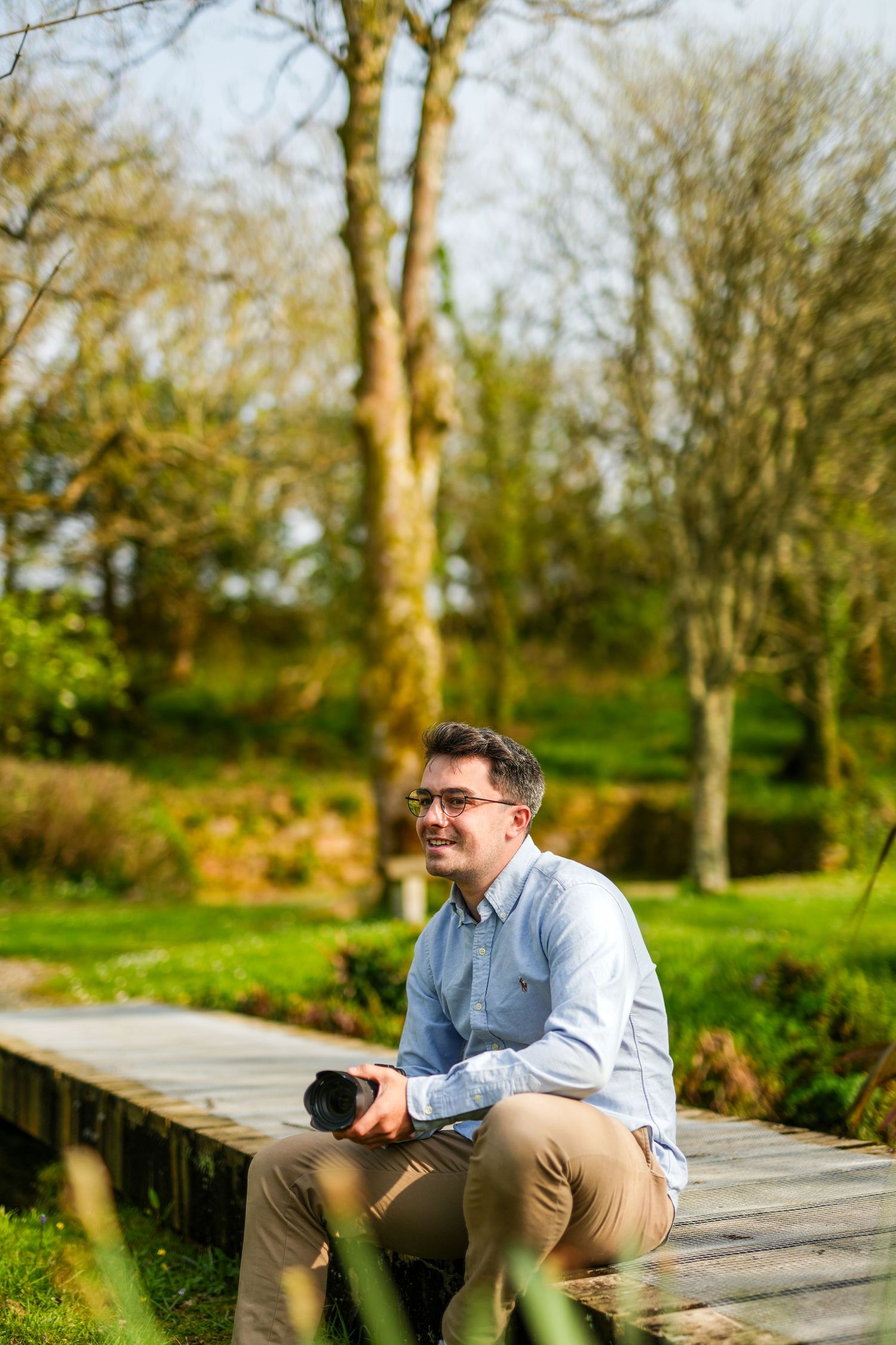 Photographer sitting on wooden boardwalk in nature setting surrounded by trees and greenery on sunny day.