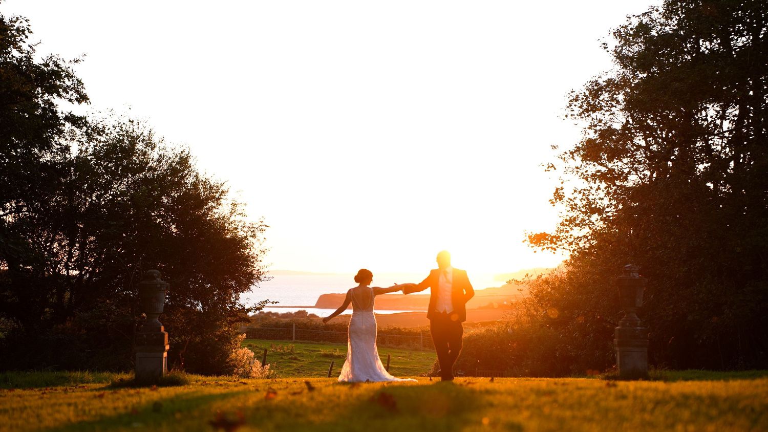 A couple holds hands while walking in a golden sunset through a scenic outdoor field surrounded by trees.