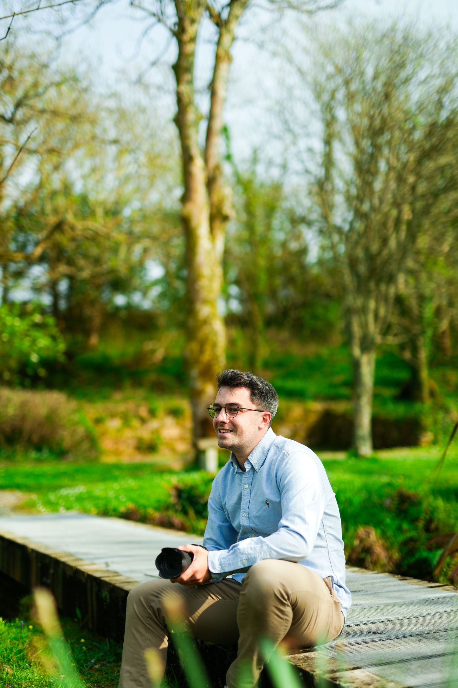 Person sitting on wooden walkway in nature setting wearing blue shirt and khakis on sunny spring day.