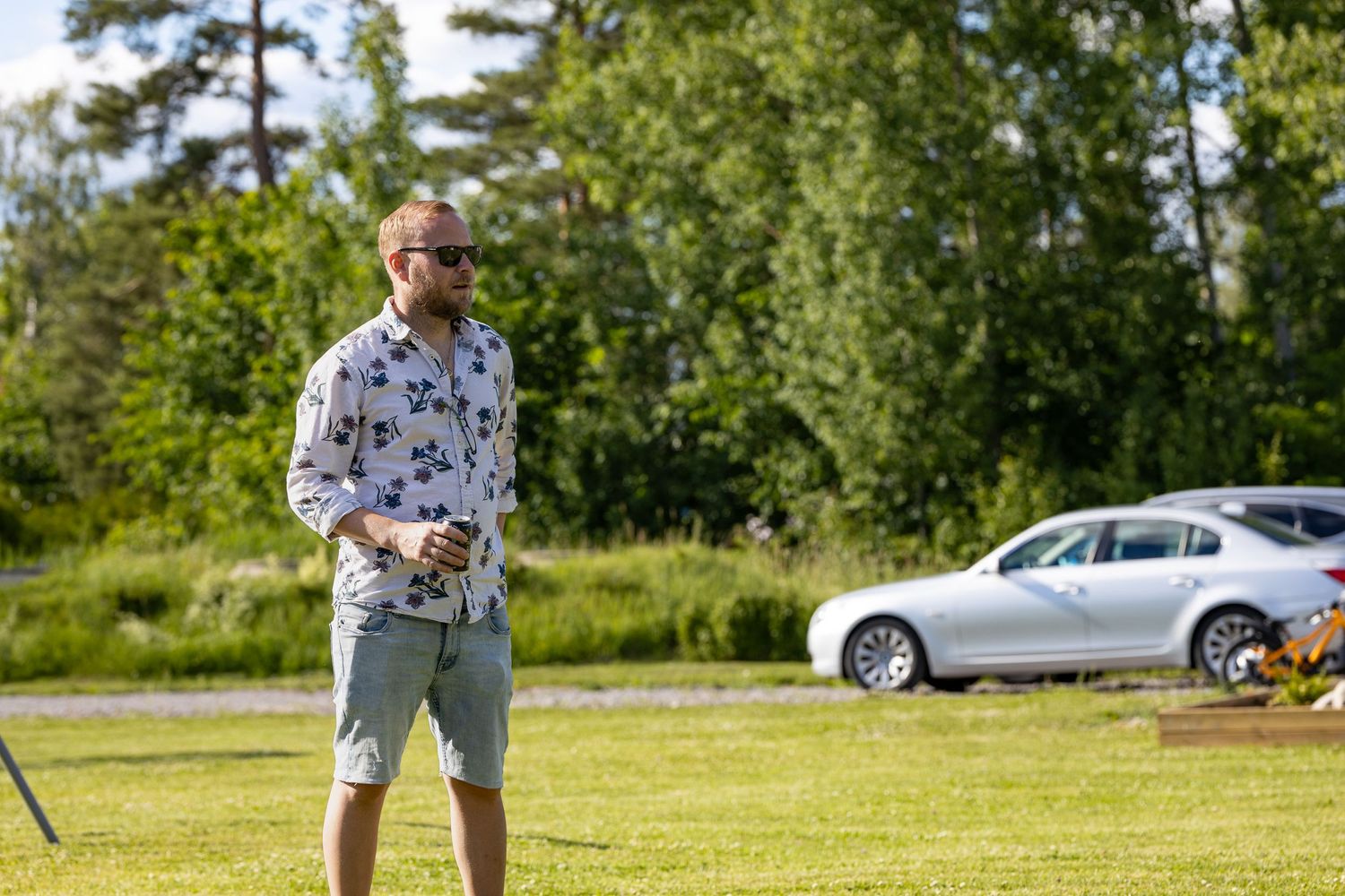 En person iklädd mönstrad skjorta och shorts står på en gräsmatta med parkerade bilar i bakgrunden.