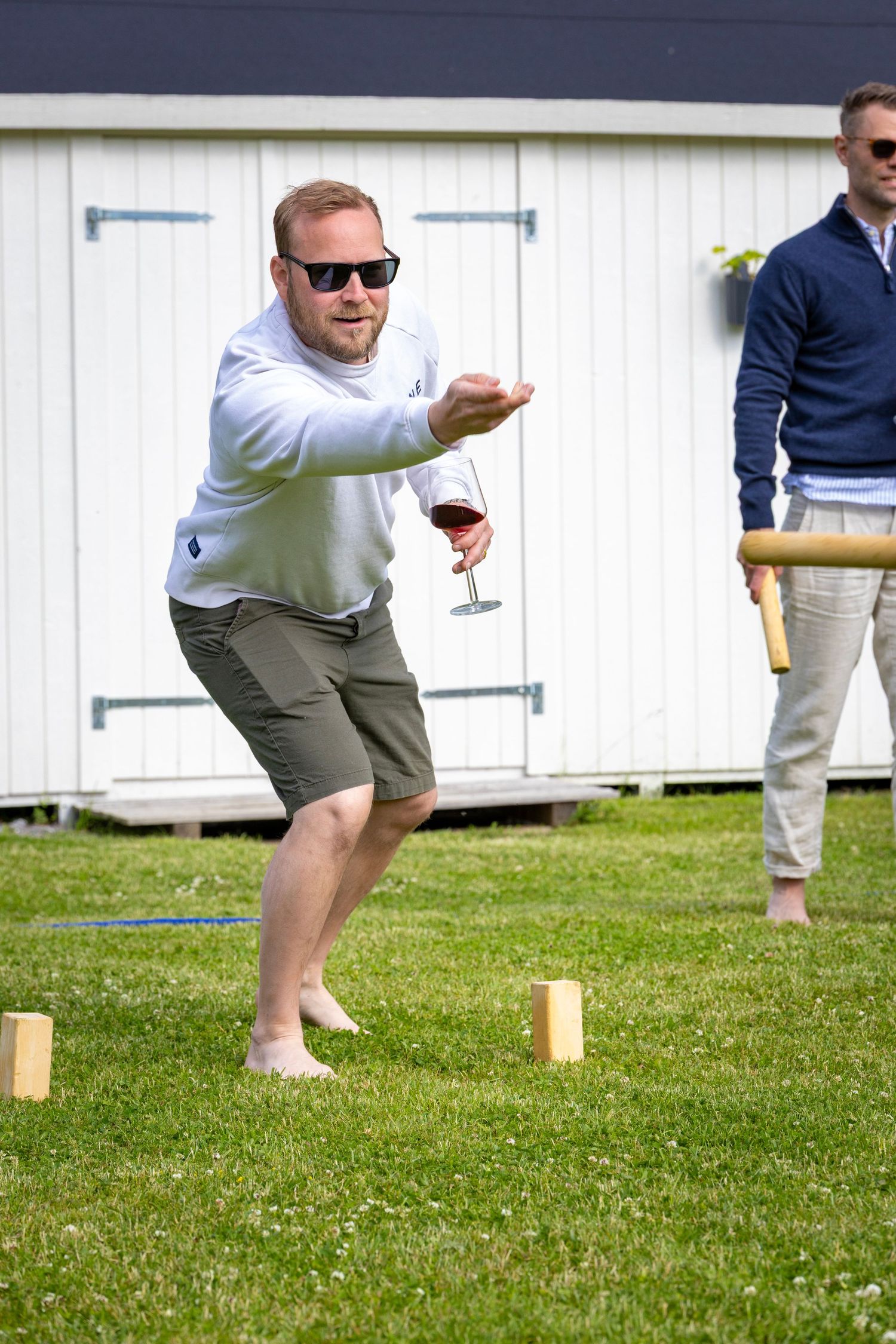 En spelare med solglasögon kastar en kubb under ett kubb spel på gräsmattan.