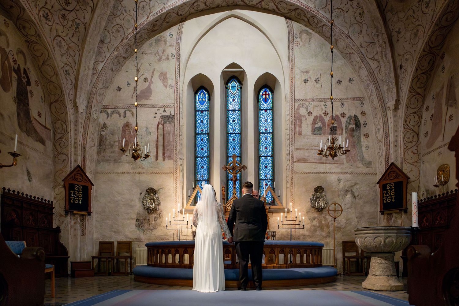 Ett brudpar står vid altaret framför fantastiska blå glasmålningar i en historisk kyrka.