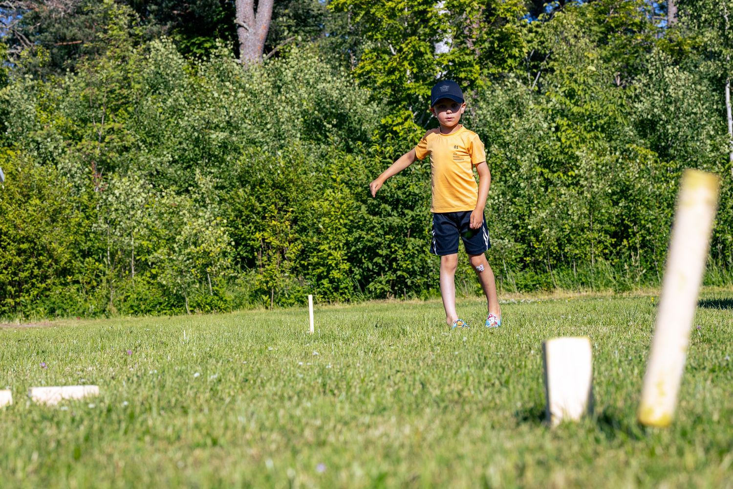 Ett barn i en gul tröja som spelar ett kubb på en solig dag.
