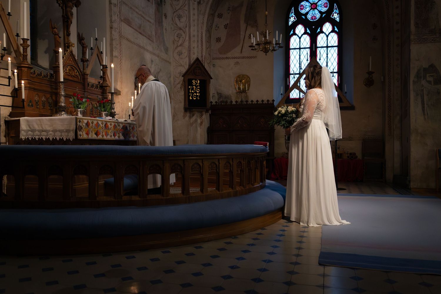 En brudklänning står i tyst reflektion framför ett utsmyckat kyrkaltare med glödande blå fönster.