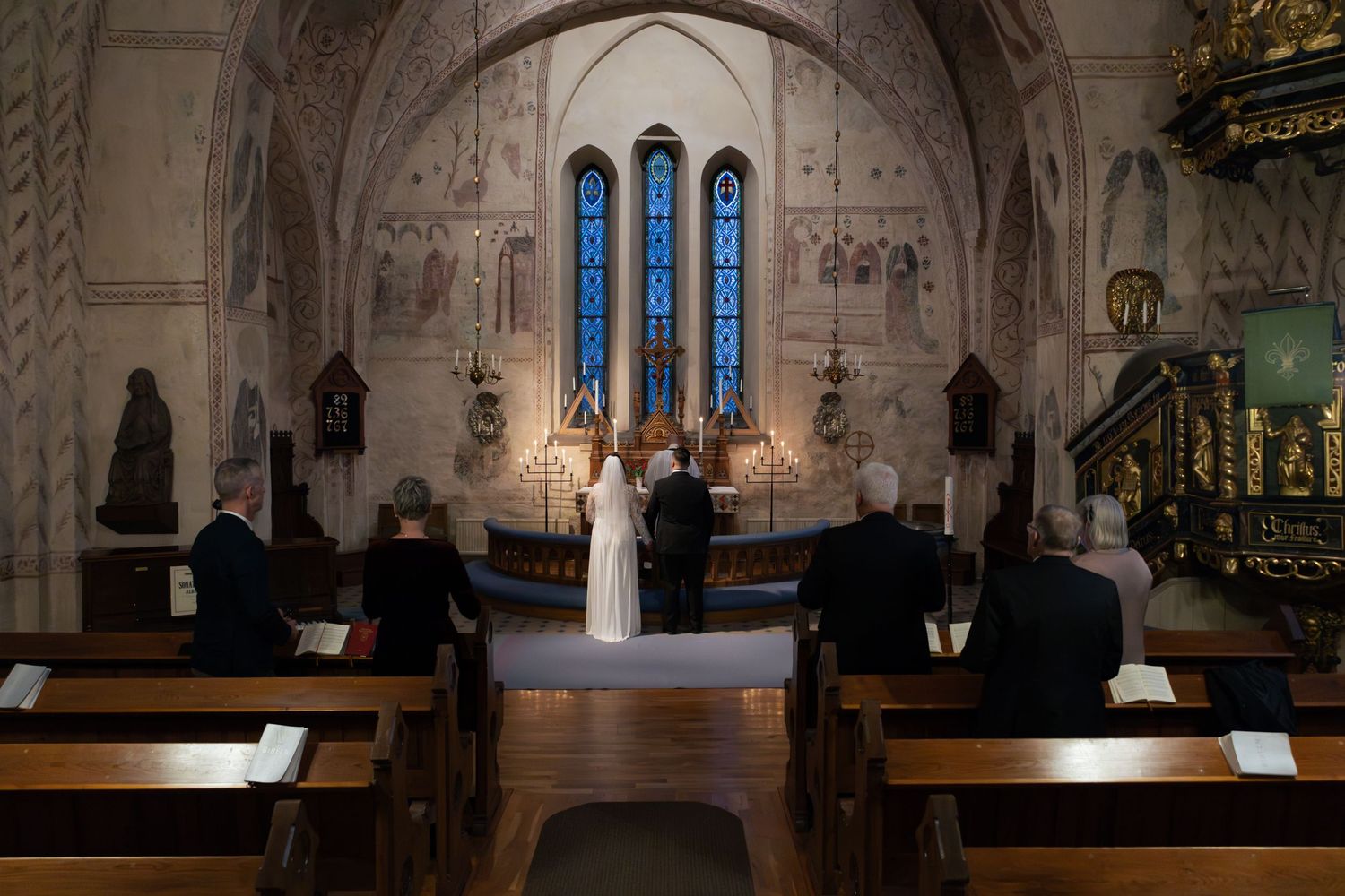 En liten kyrklig vigselceremoni med vackra blå glasmålningar och träbänkar.