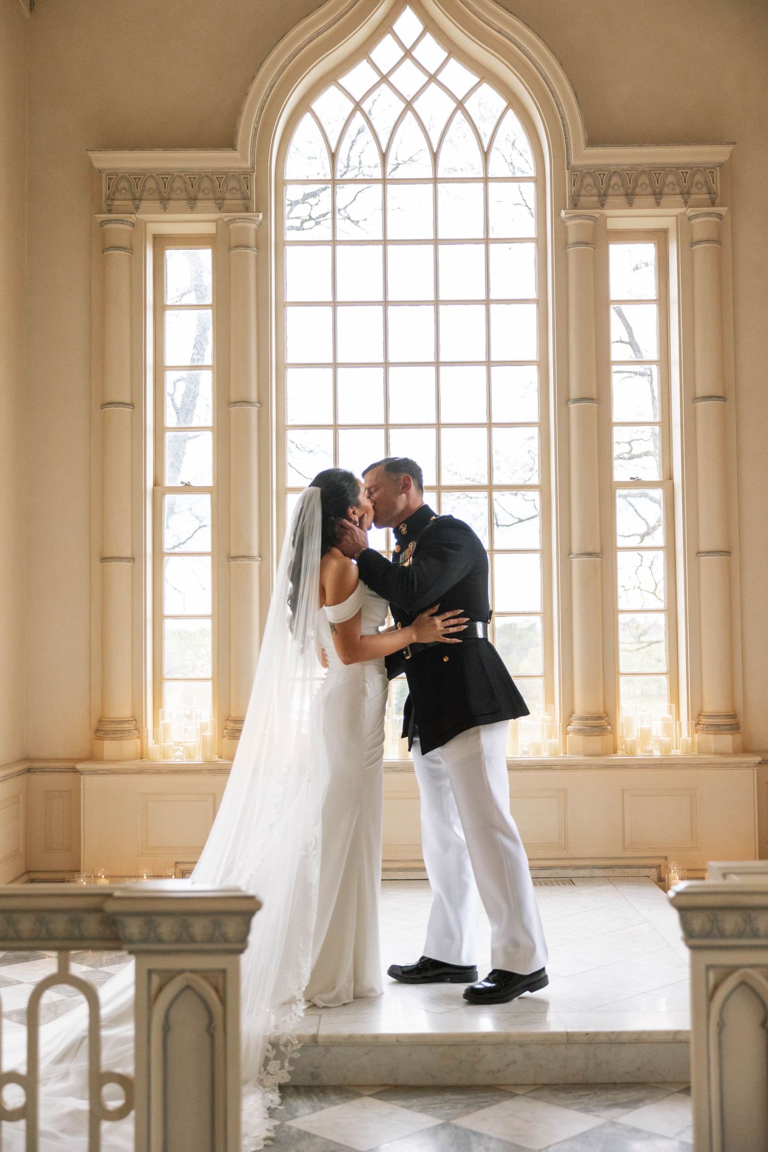 A wedding couple shares a romantic moment in front of a grand Gothic arched window in an elegant chapel interior.