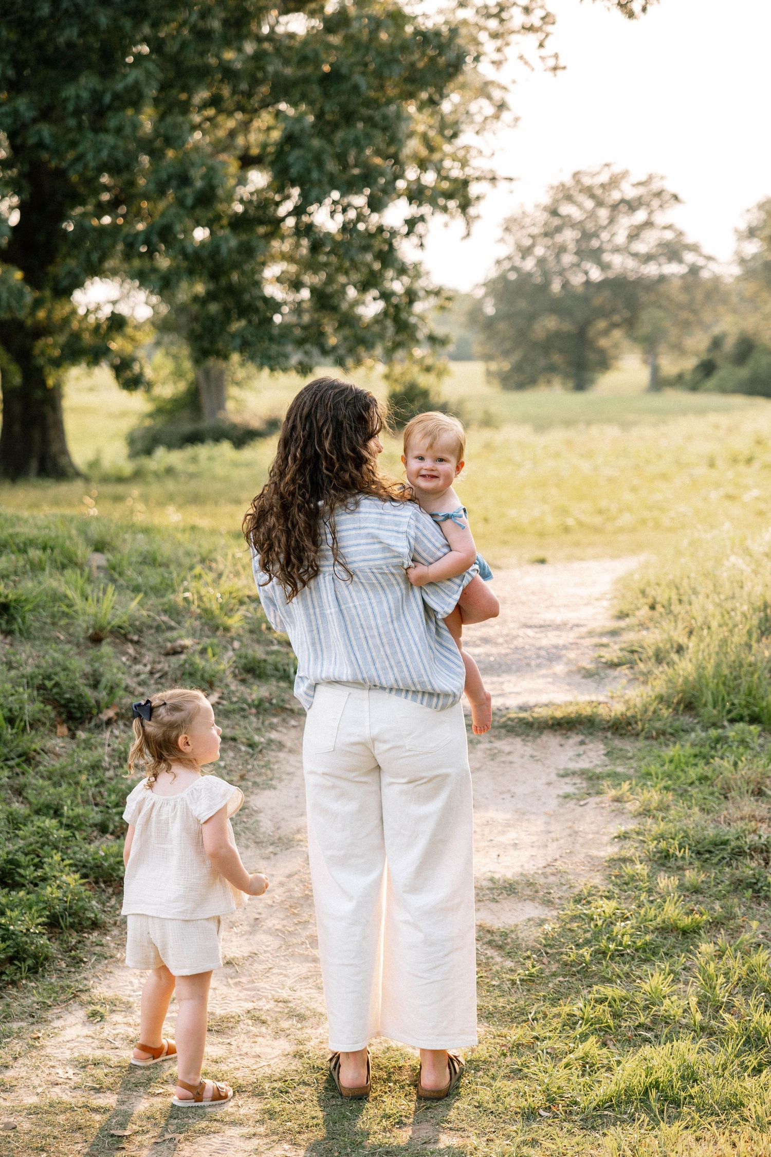 A family walks together on a sunlit dirt path through a serene meadow on a warm summer evening.