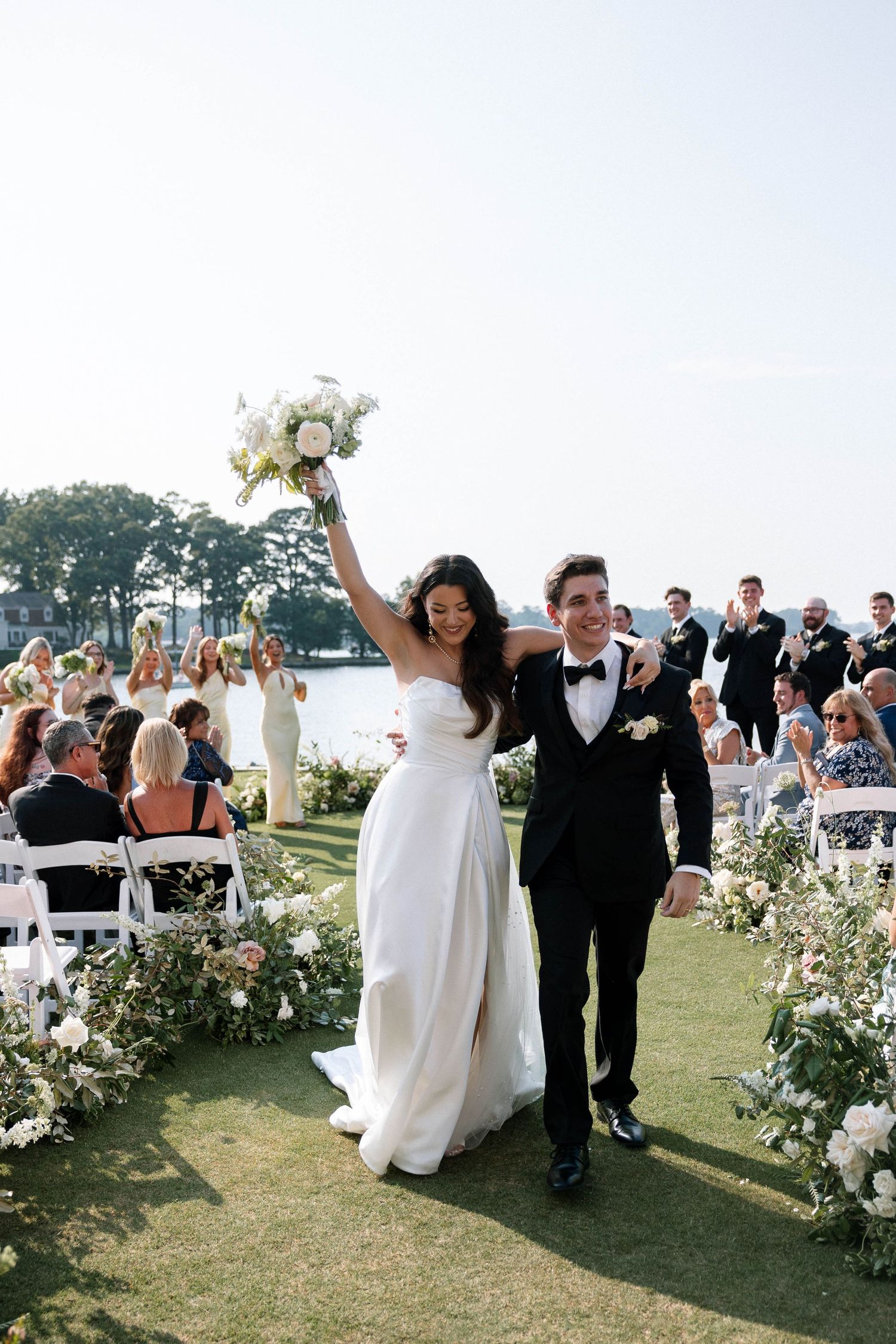 Newlyweds joyfully walk down the aisle at a scenic outdoor wedding ceremony decorated with white flowers by the water.