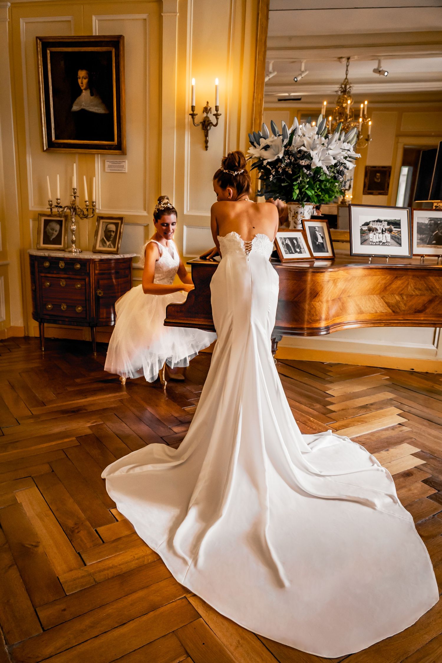 Elegant bride in strapless mermaid gown with long train stands in opulent room with chandelier and ornate decor.