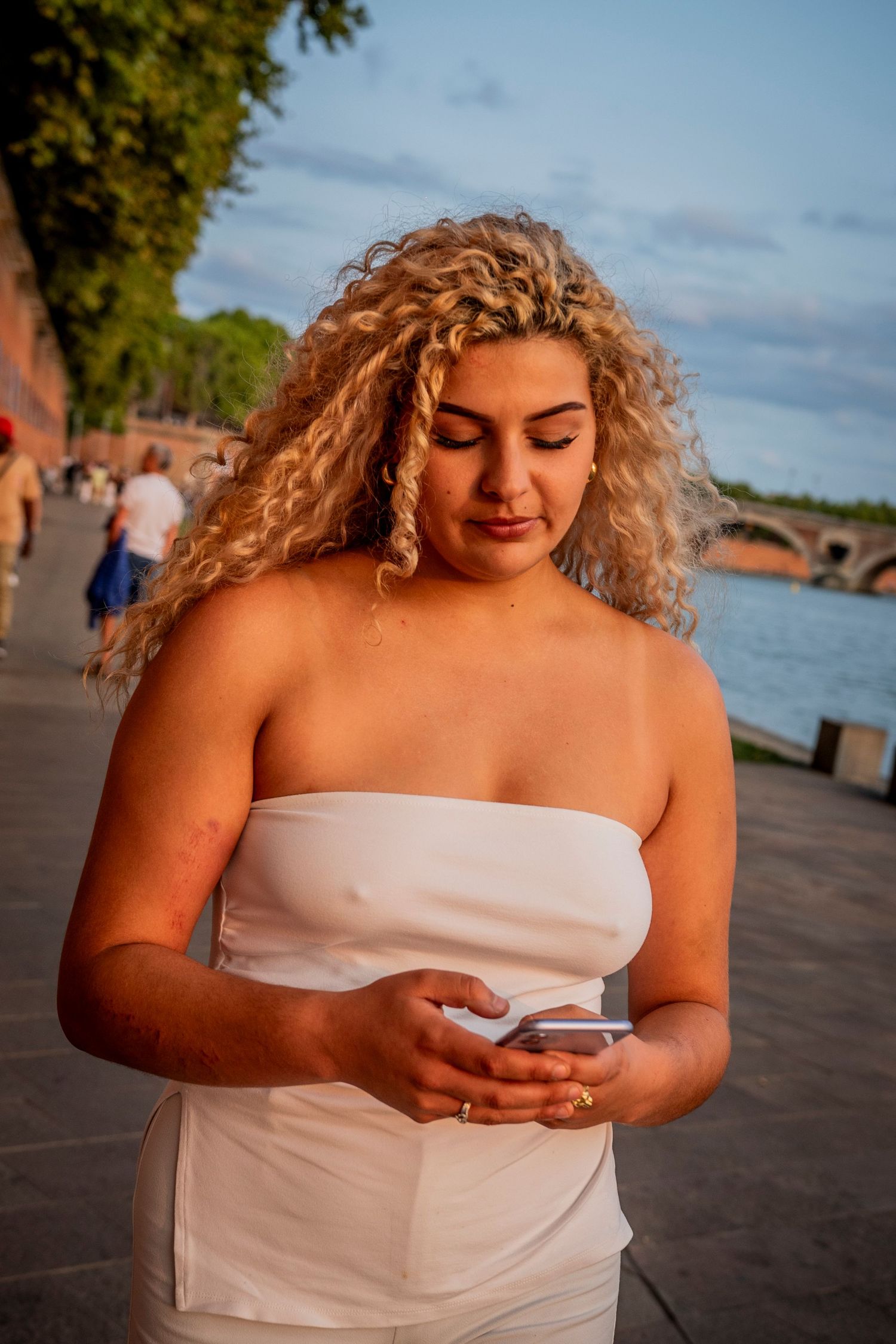 A series of photos shows someone in a white strapless dress looking down at a mobile phone on a waterfront promenade.