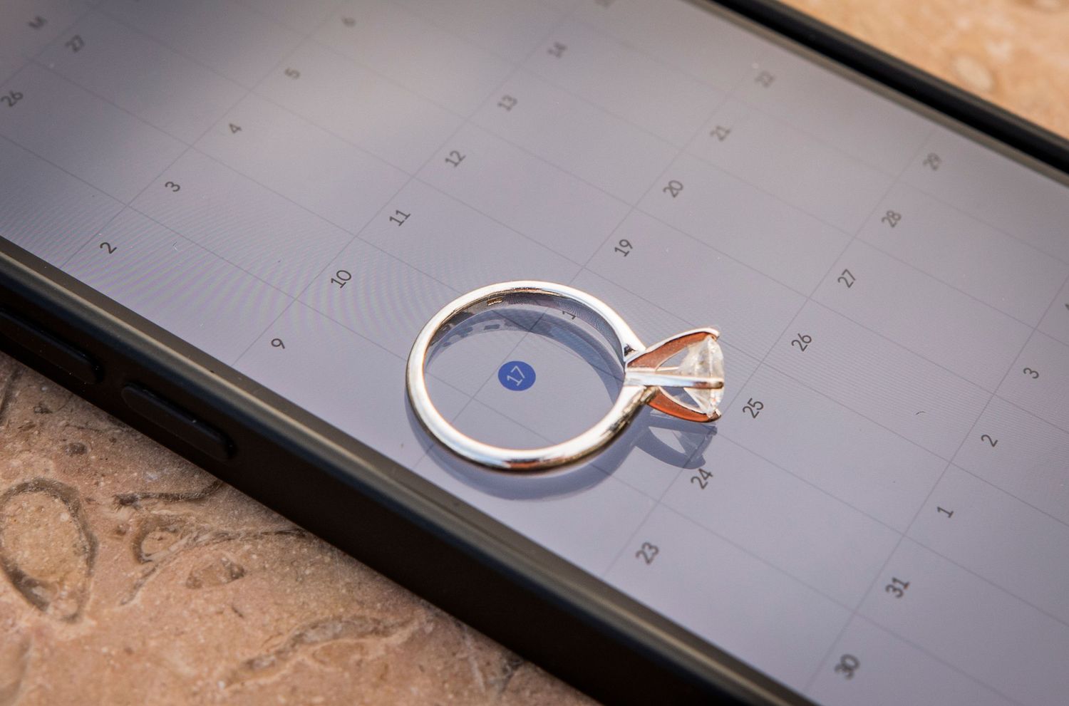 A delicate rose gold and silver engagement ring sits on top of a dark smartphone screen displaying a calendar layout.