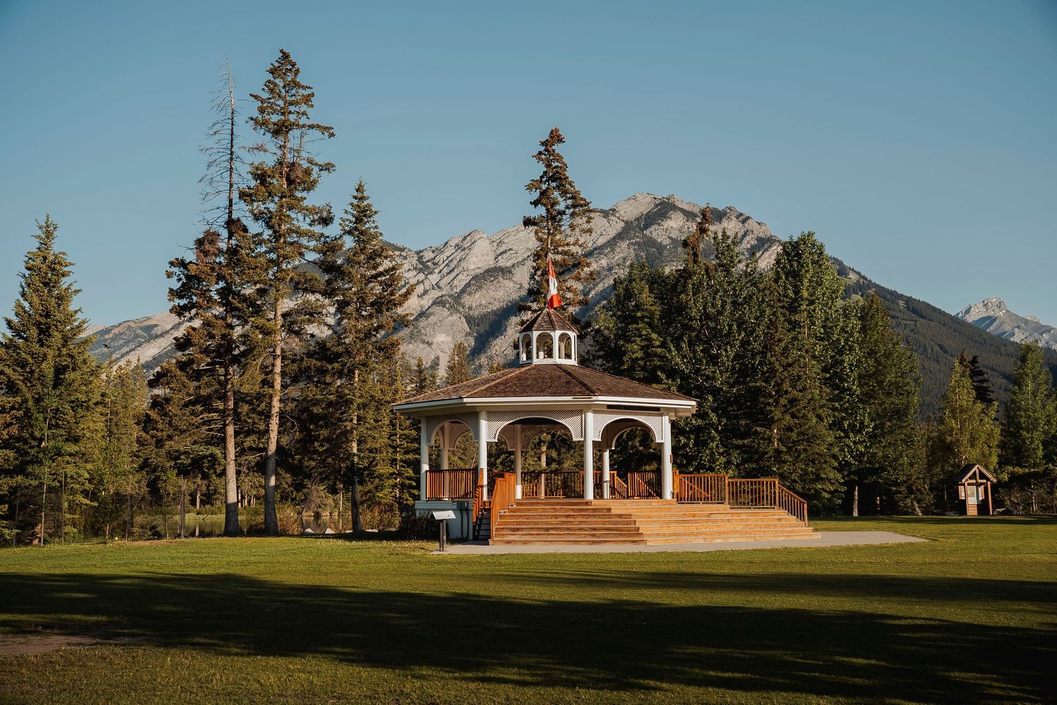 A rustic wooden gazebo stands in front of snow-capped mountain peaks at Banff National Park, surrounded by evergreen trees.