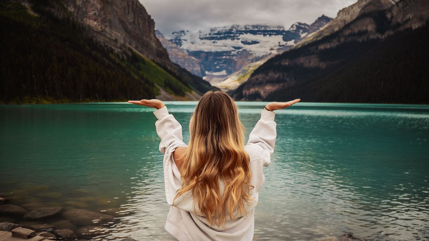 Person with long hair overlooking turquoise Lake Louise with snow-capped mountains in Banff National Park, Canada.