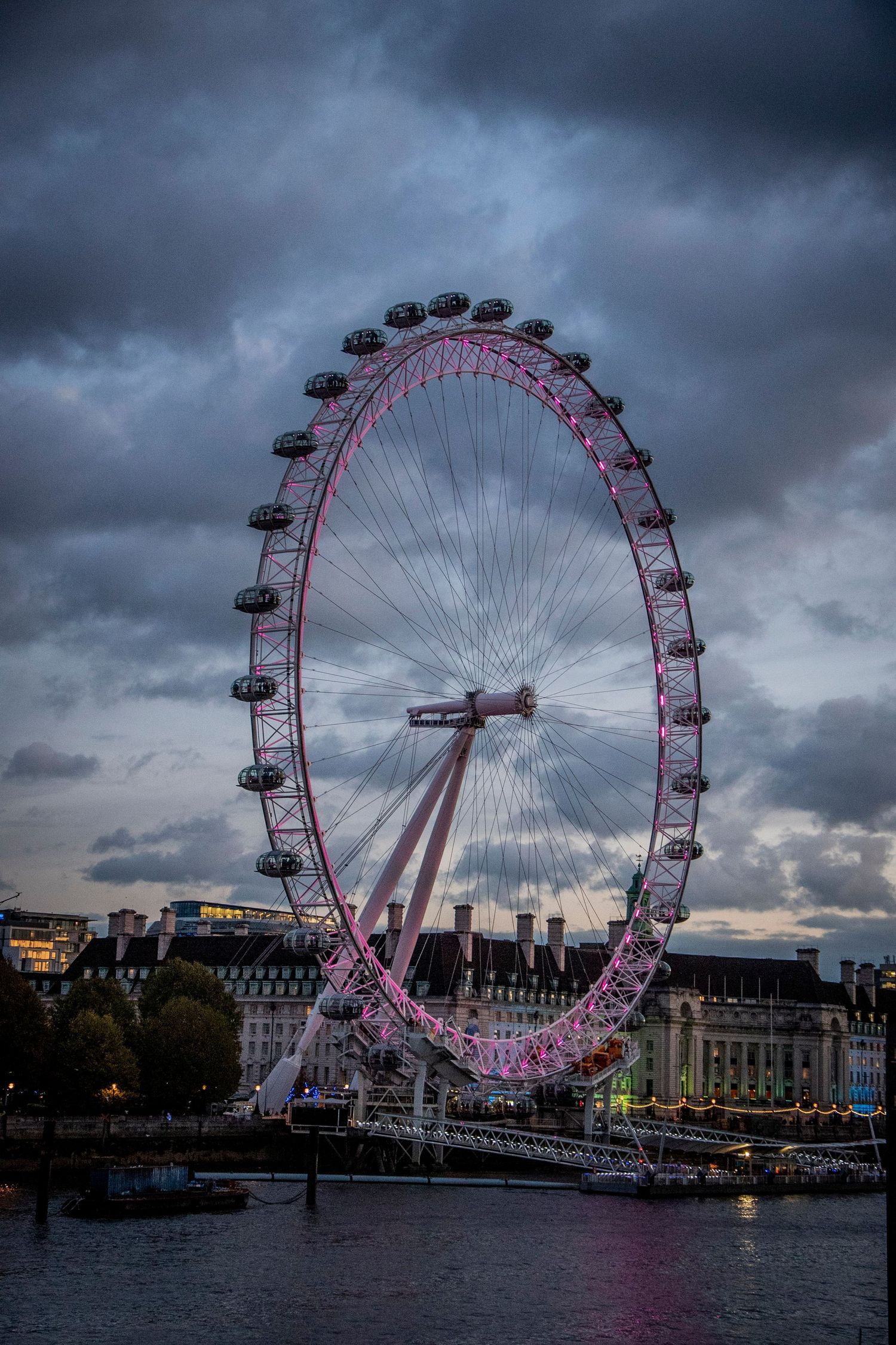 The London Eye Ferris wheel illuminated in pink against dramatic stormy clouds over the River Thames at dusk.
