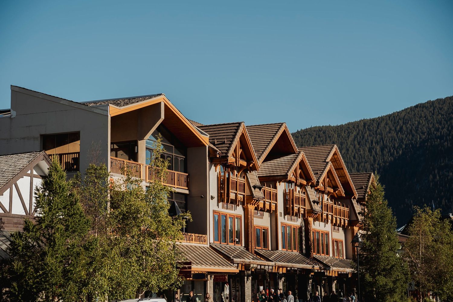 Alpine-style lodge with wooden balconies and gabled roofs nestled against forested mountain backdrop under blue sky.