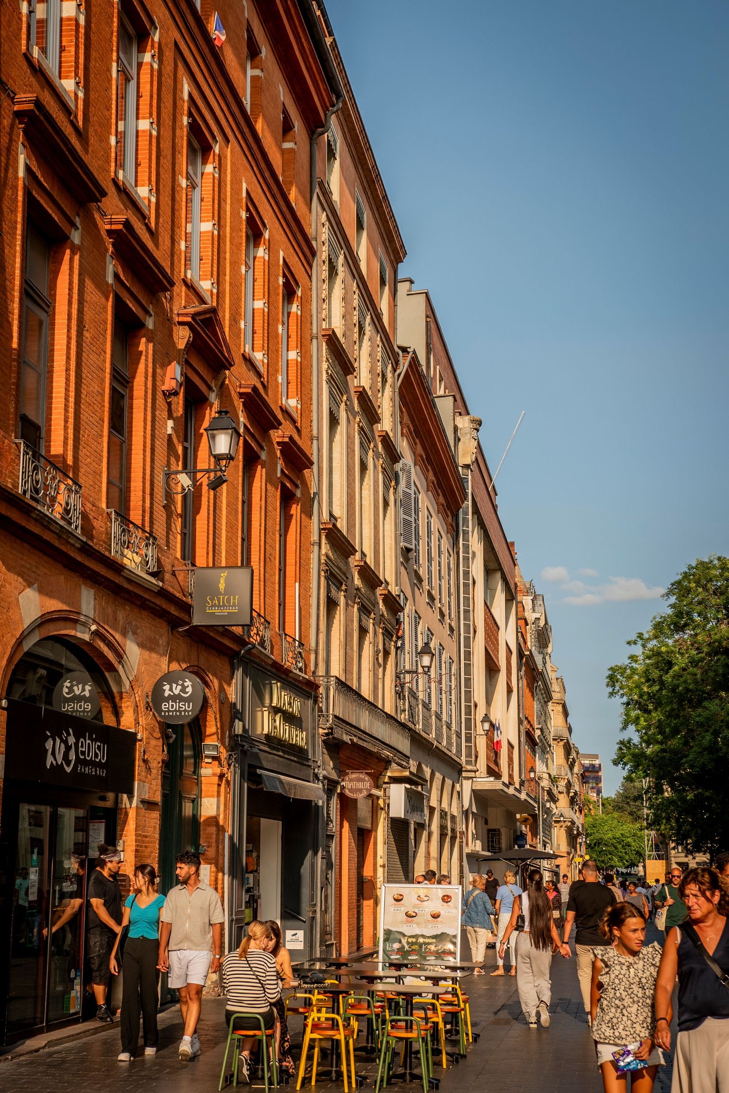 People walking on a charming European street lined with historic brick buildings and outdoor cafe seating on a sunny day.