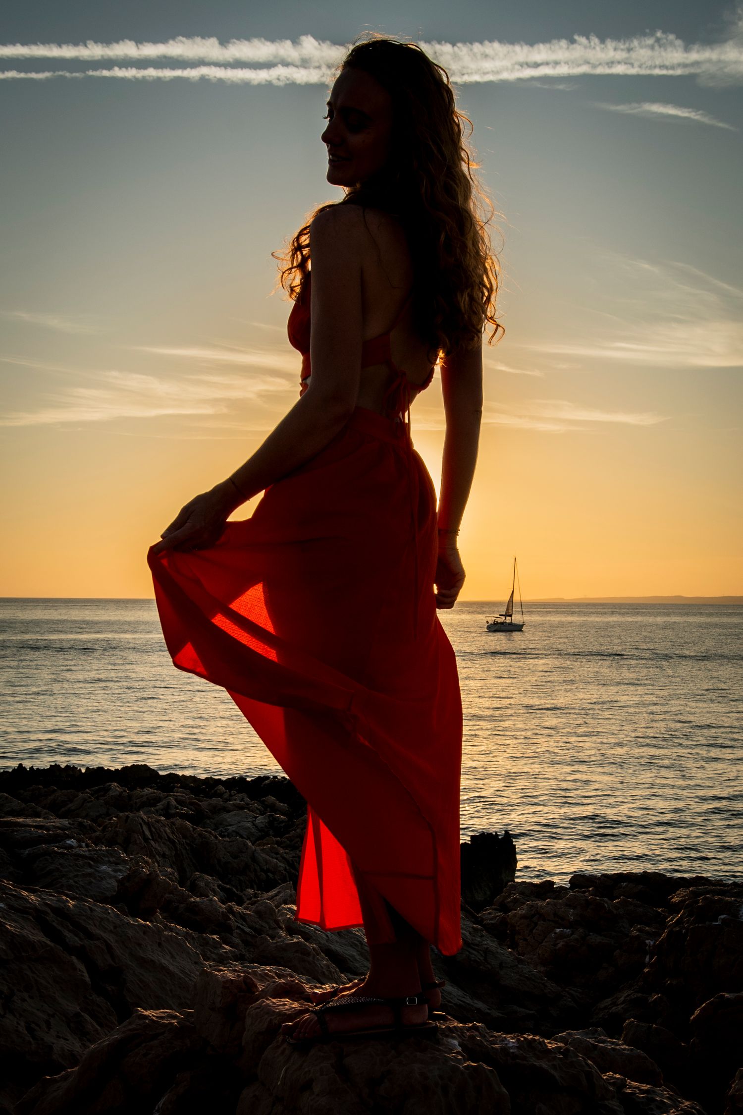 Silhouette of someone in a flowing red dress standing on rocky coastline at sunset with sailboat in distance on ocean horizon.