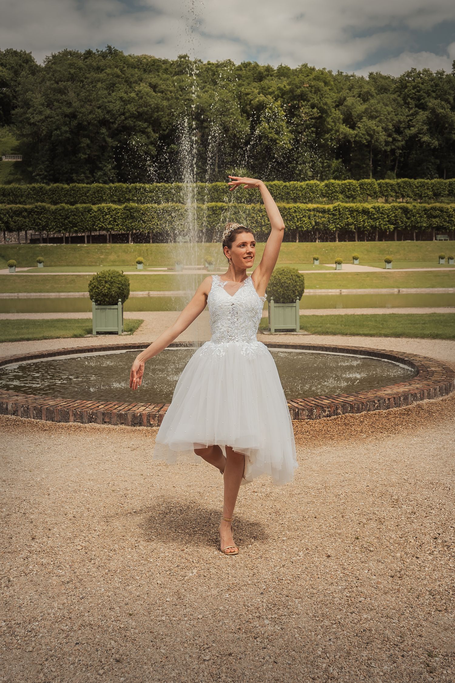 A dancer in a white tulle dress poses gracefully near a fountain in a formal garden setting.