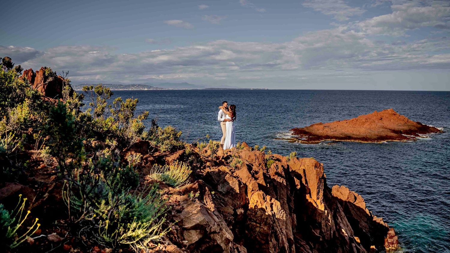 A couple stands on red coastal rocks overlooking turquoise waters and dramatic ocean views at sunset.